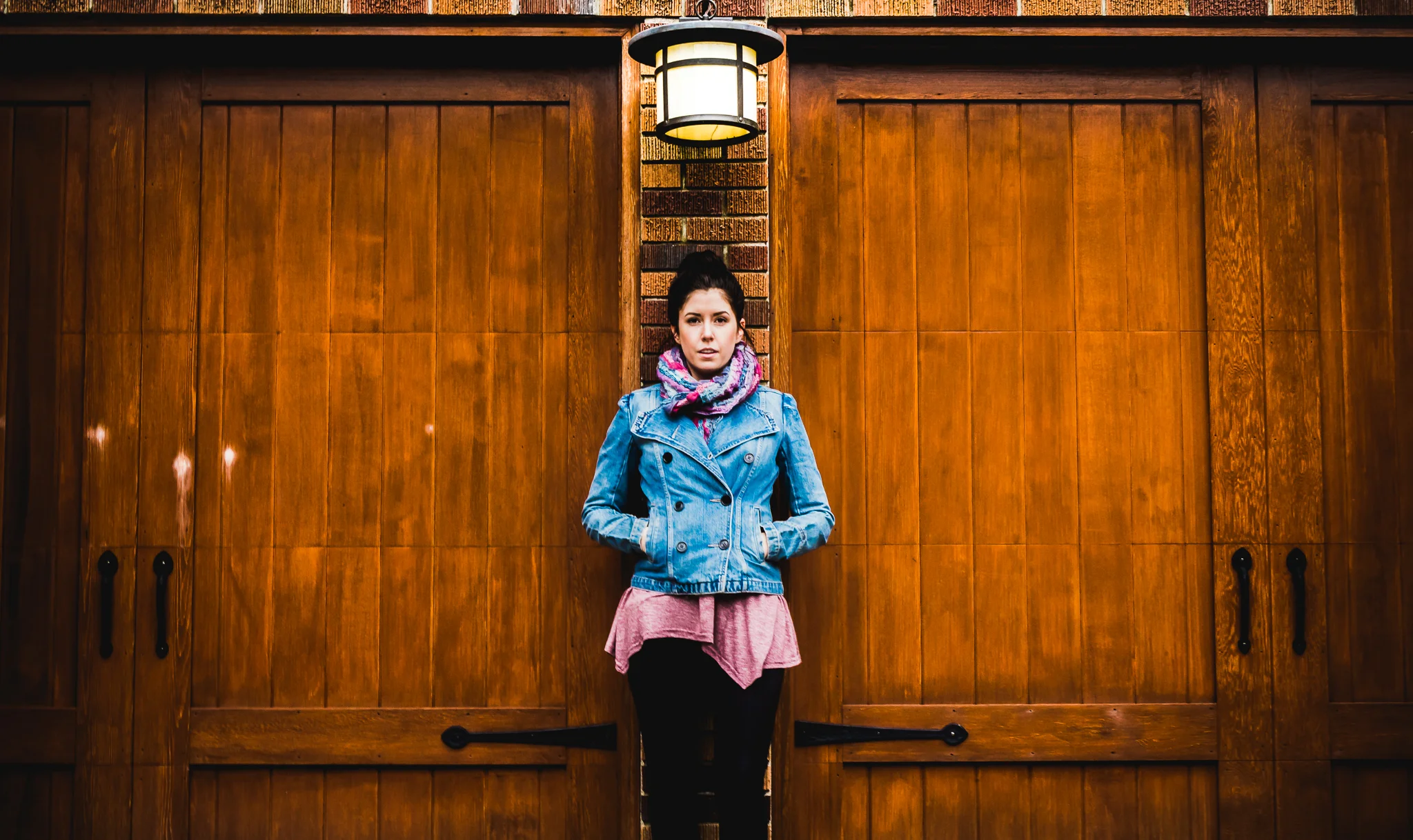 A woman stands in front of a wooden garage door, wearing a denim jacket, pink shirt, and a colorful scarf.