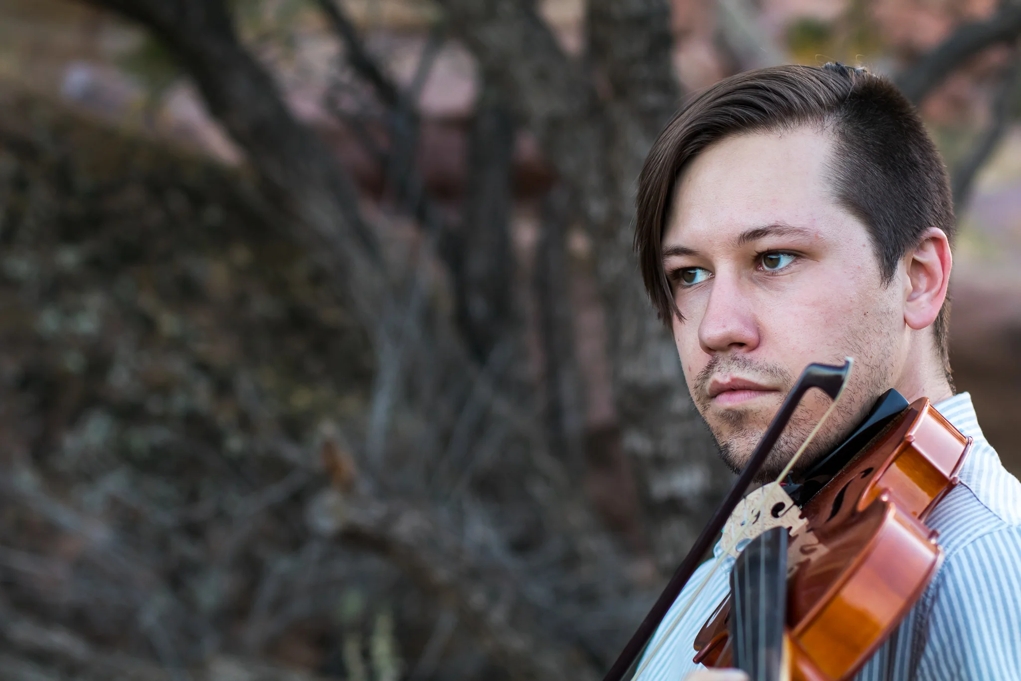 Young man with dark hair playing a violin outdoors with trees in the background.