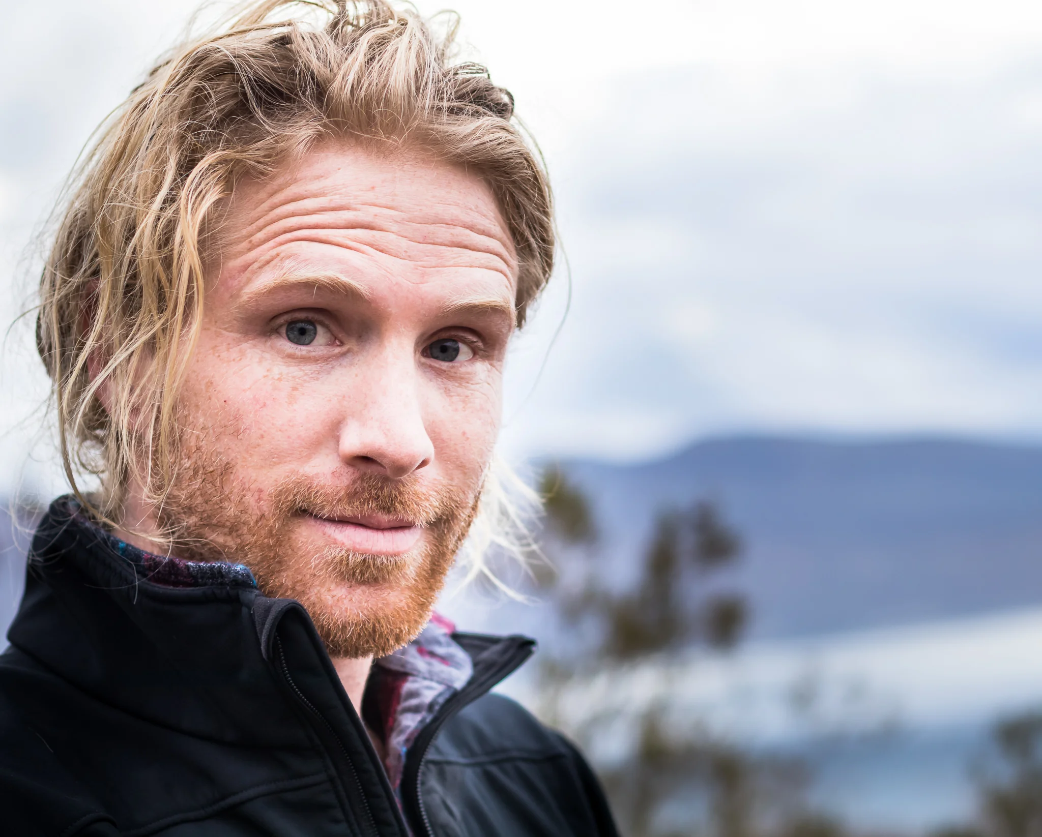 Close-up of a man with long blonde hair, a beard, wearing a black jacket, outdoors with a cloudy sky and mountains in the background.