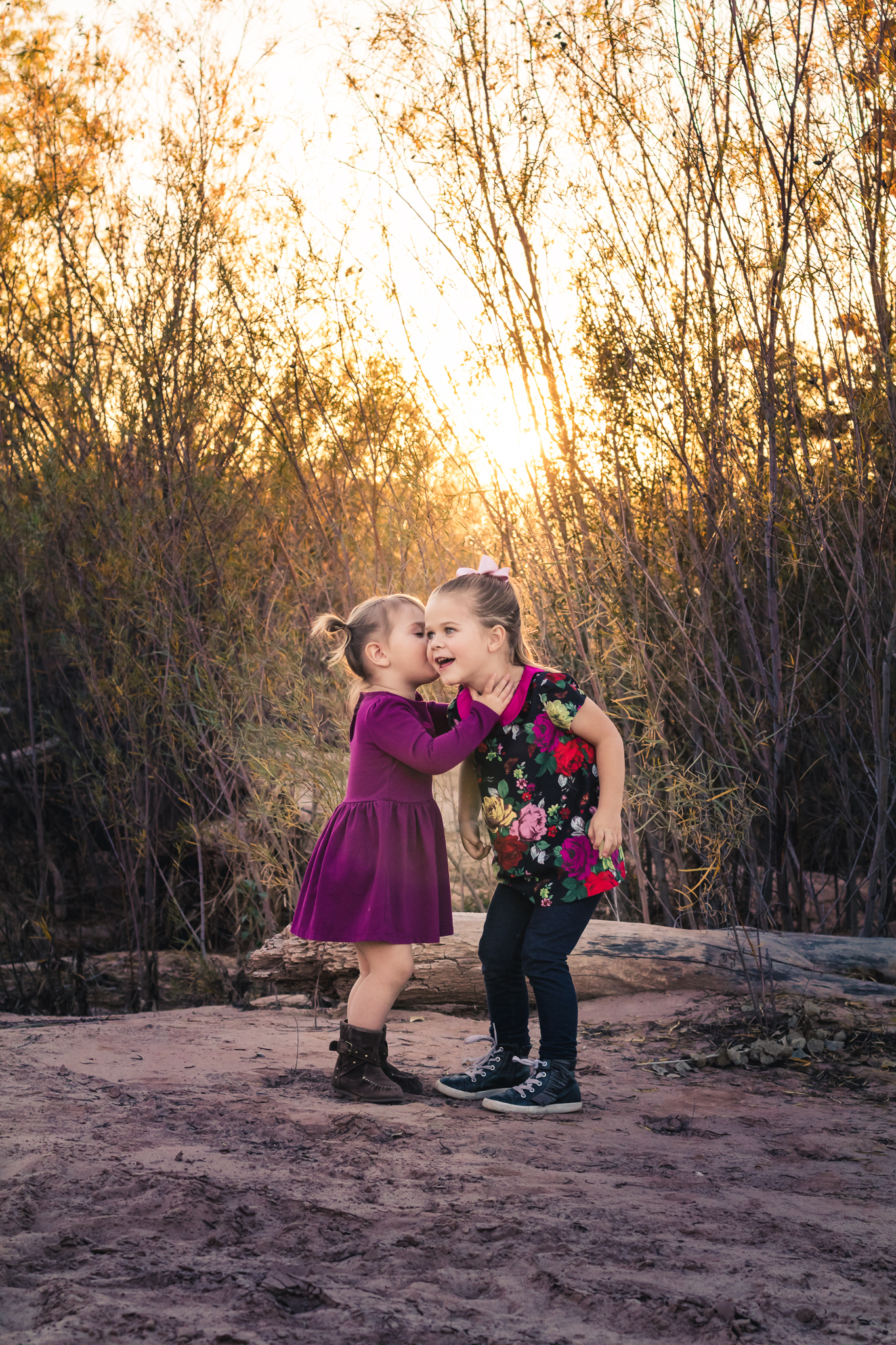 Two young girls sharing a secret or whispering to each other outdoors at sunset, surrounded by trees and fallen logs.