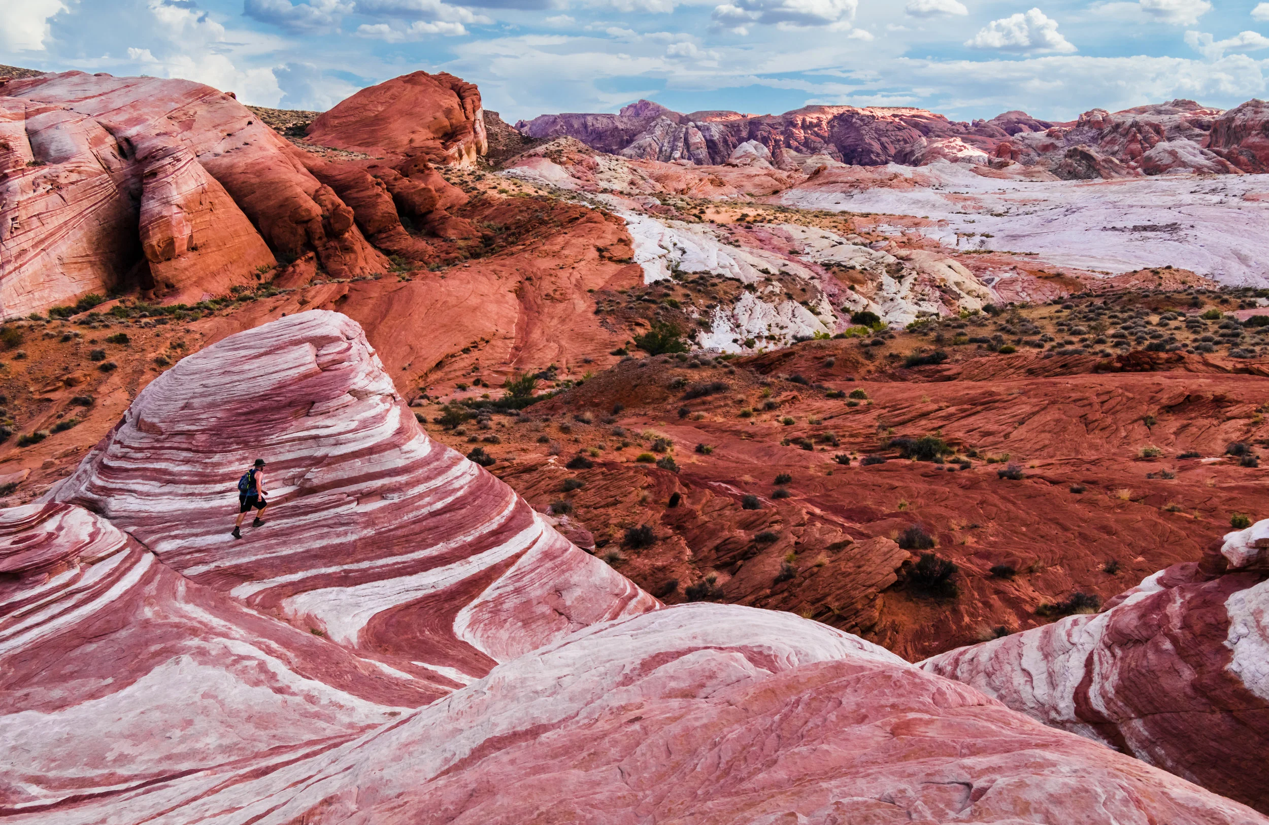 A person with a backpack hiking on a colorful, striped rock formation in a desert landscape with red, white, and pink hues, surrounded by rugged terrain and distant mountains under a partly cloudy sky.