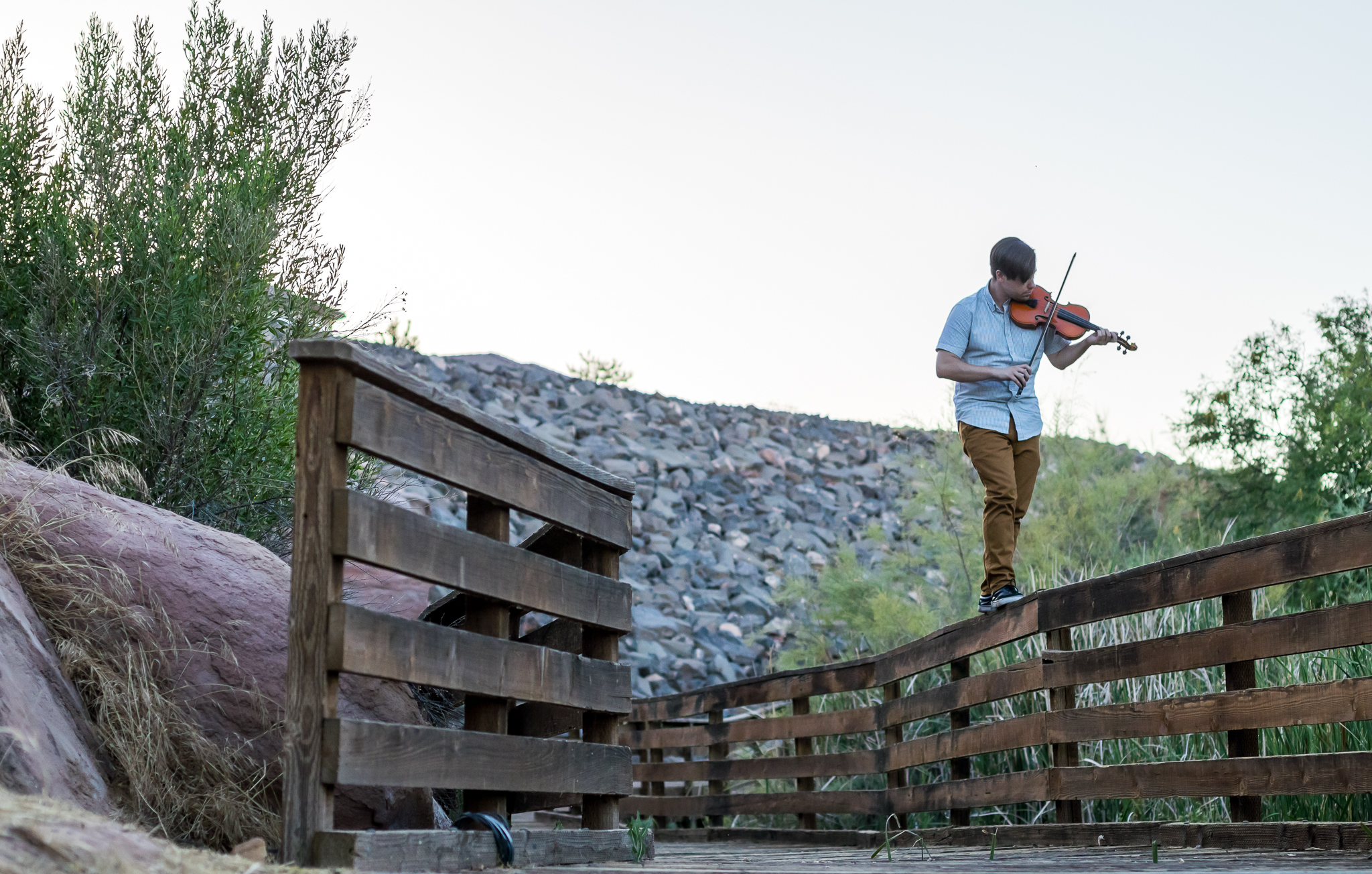 A young man in a light blue shirt and brown pants playing the violin while walking on a wooden pathway outdoors, with bushes and a rocky hillside in the background.
