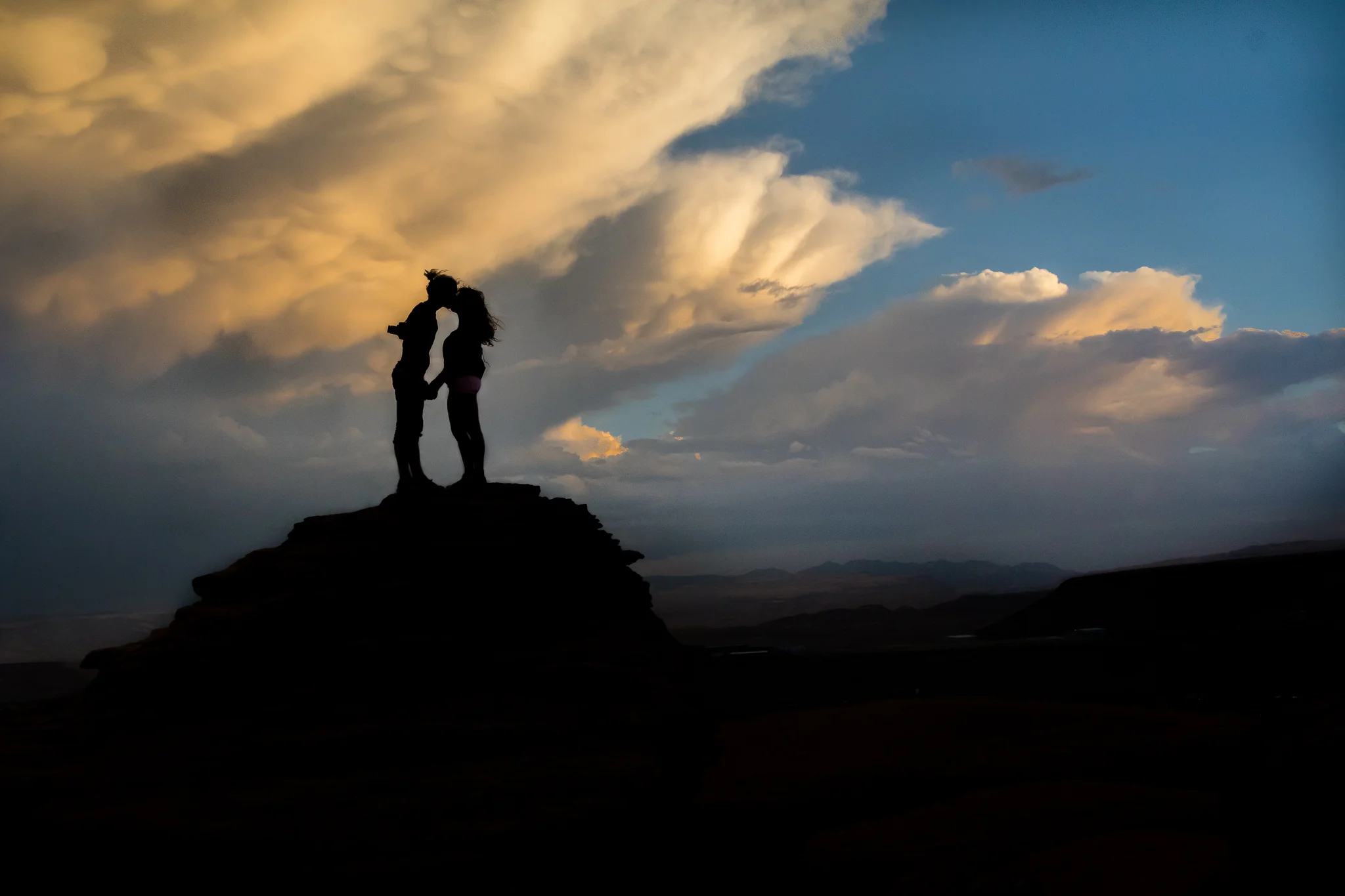 Silhouettes of two people standing close together on a rock formation at sunset with a colorful sky and clouds in the background.