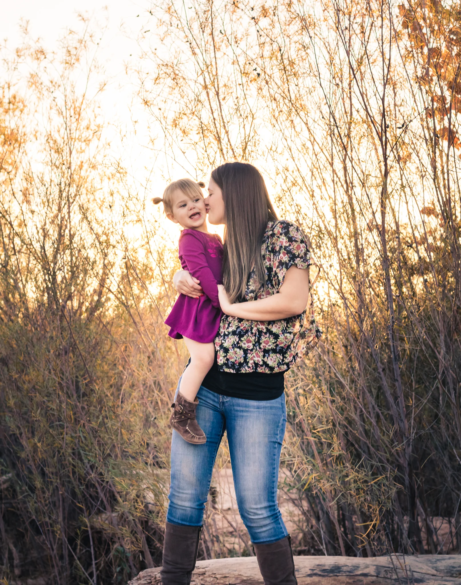 A woman holding a young girl outside during sunset, surrounded by leafless trees and bushes.