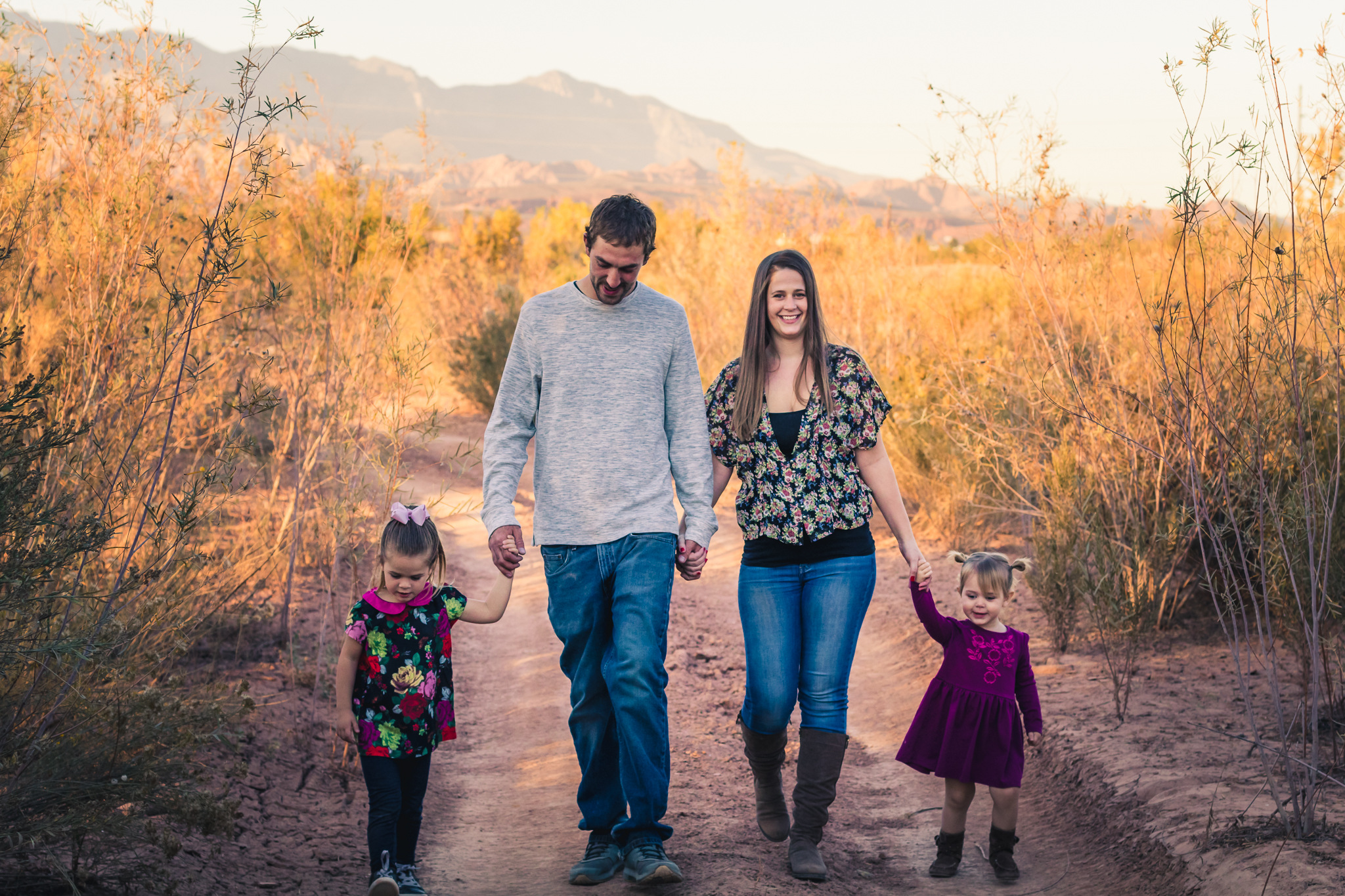 A family of four walking hand in hand on a dirt trail in a desert landscape with mountains in the background during sunset