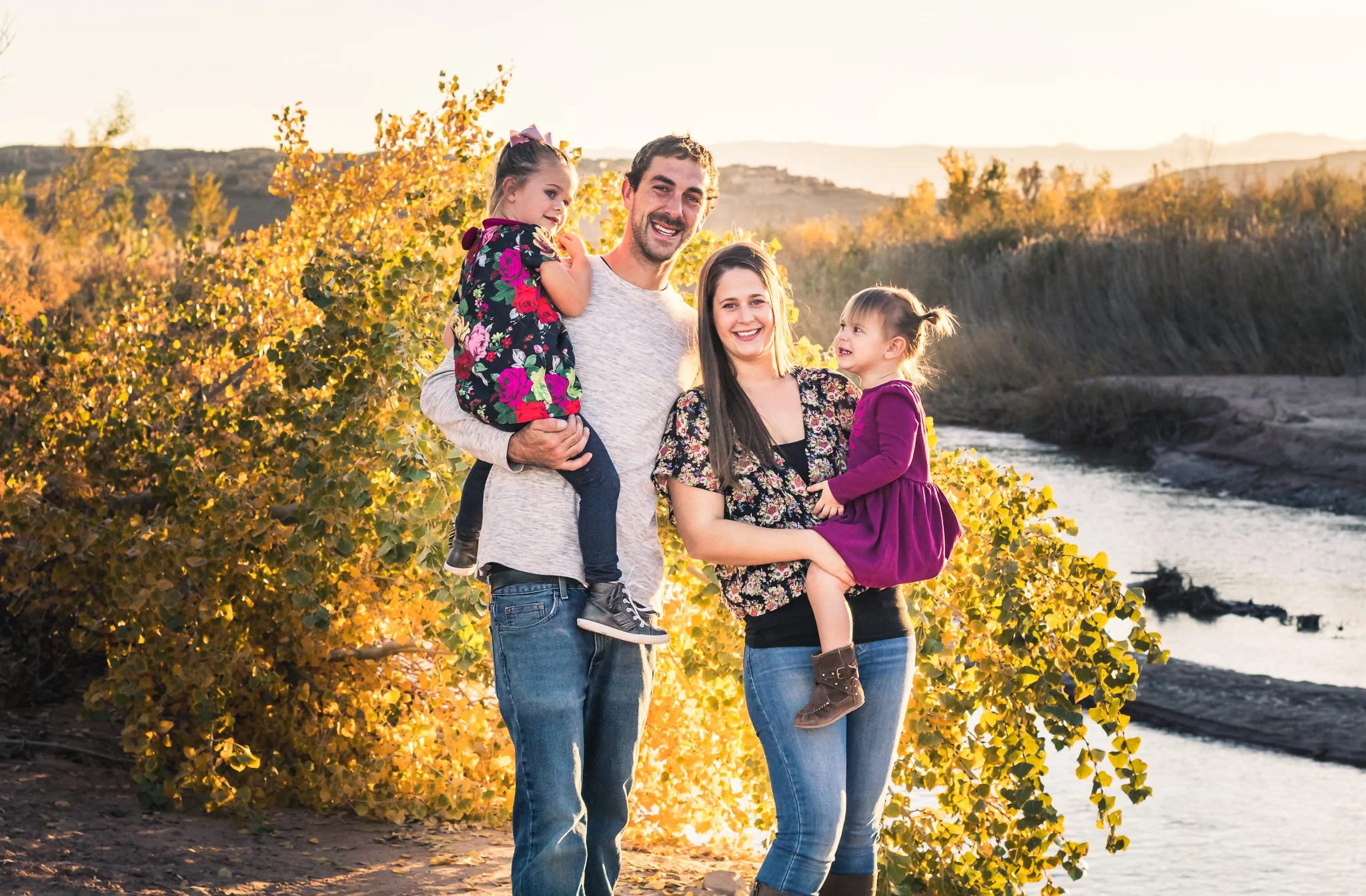 A family of four outdoors during sunset, standing near a river with autumn foliage in the background. The father is holding a young girl in a floral dress, and the mother is holding another young girl in a purple dress. All are smiling and enjoying t