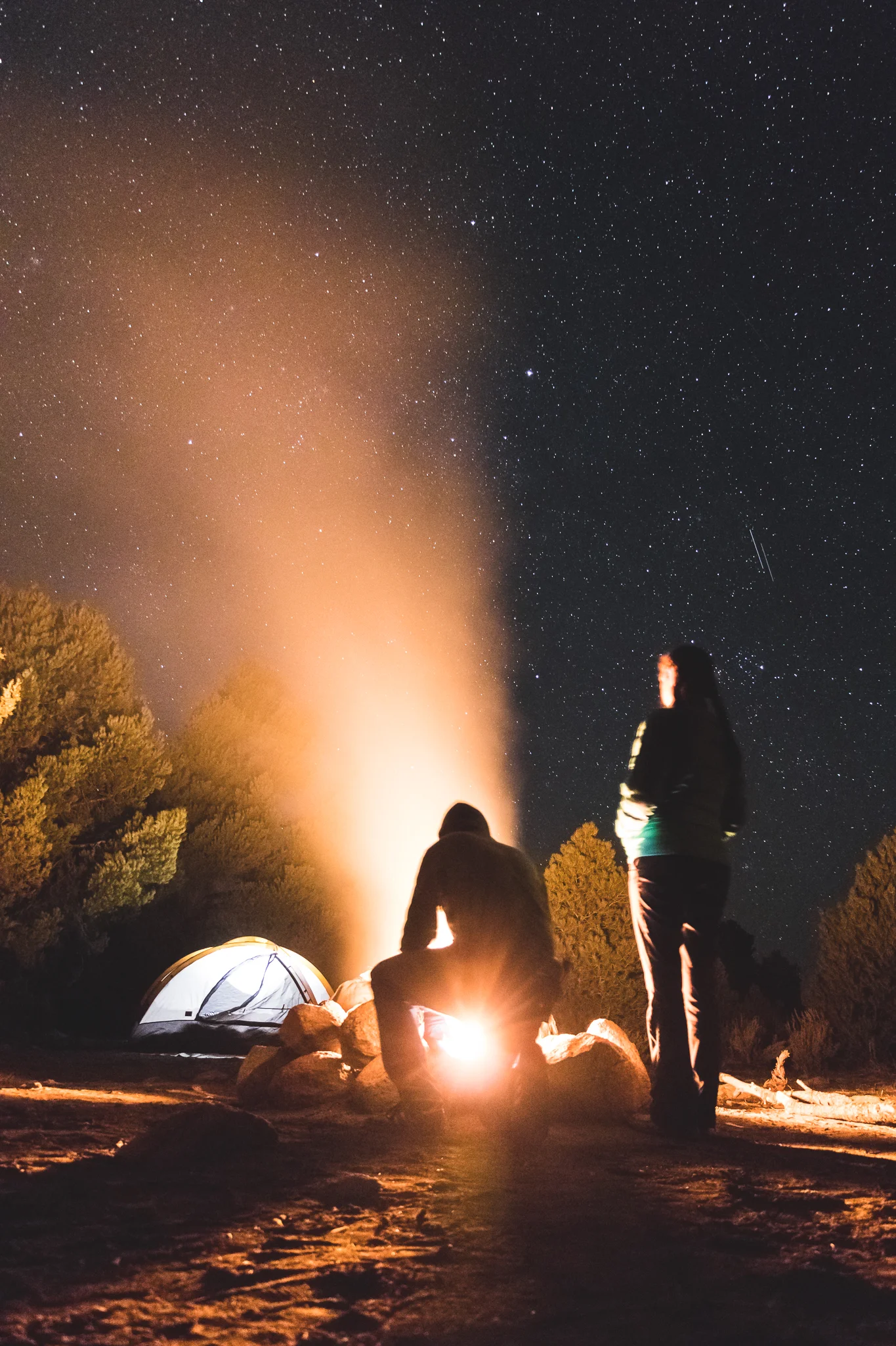 Two people camping under a starry night sky, with a tent and a campfire