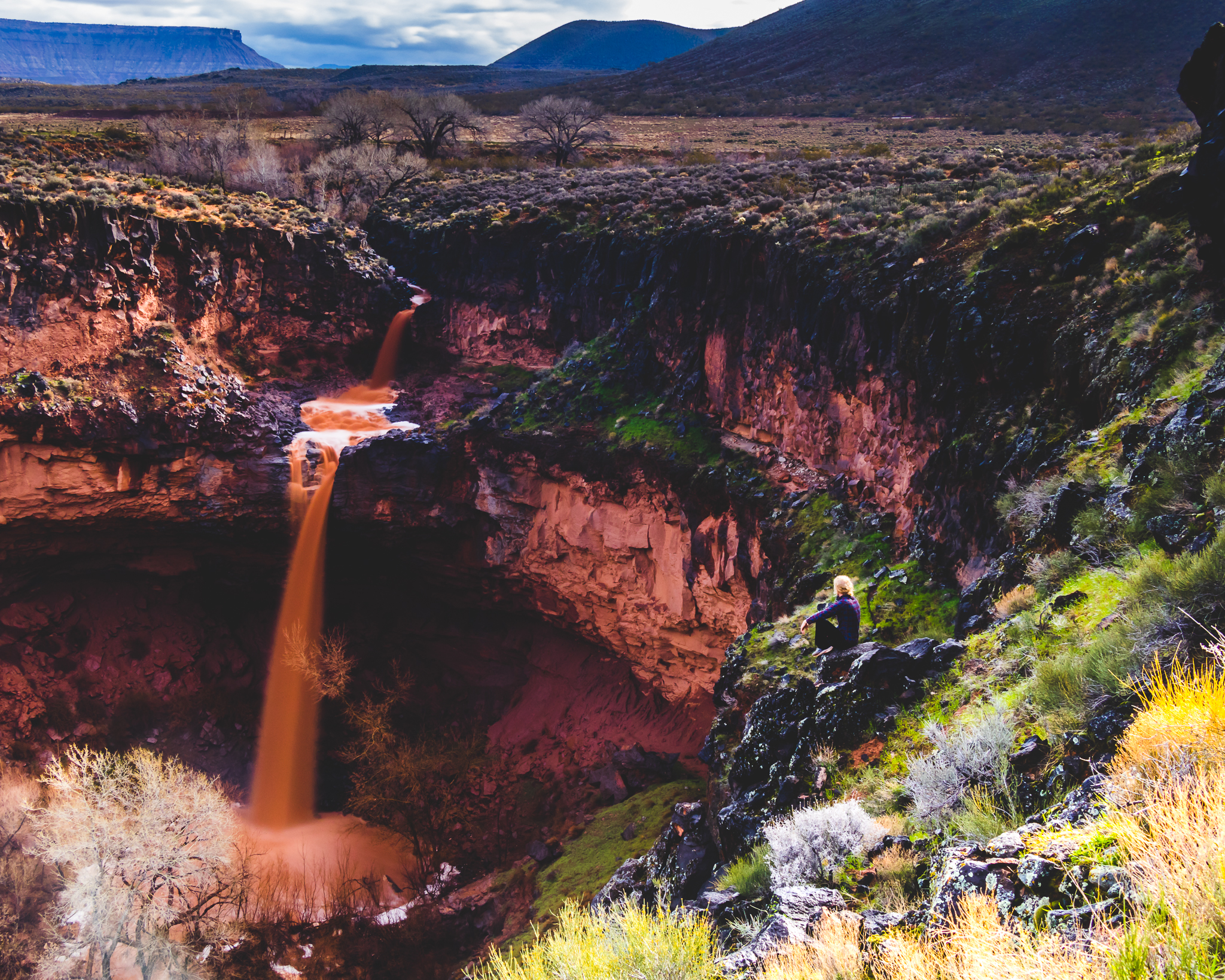 A person sitting on rocks near a canyon with a waterfall flowing into a river below, surrounded by desert vegetation and distant mountains under a partly cloudy sky.