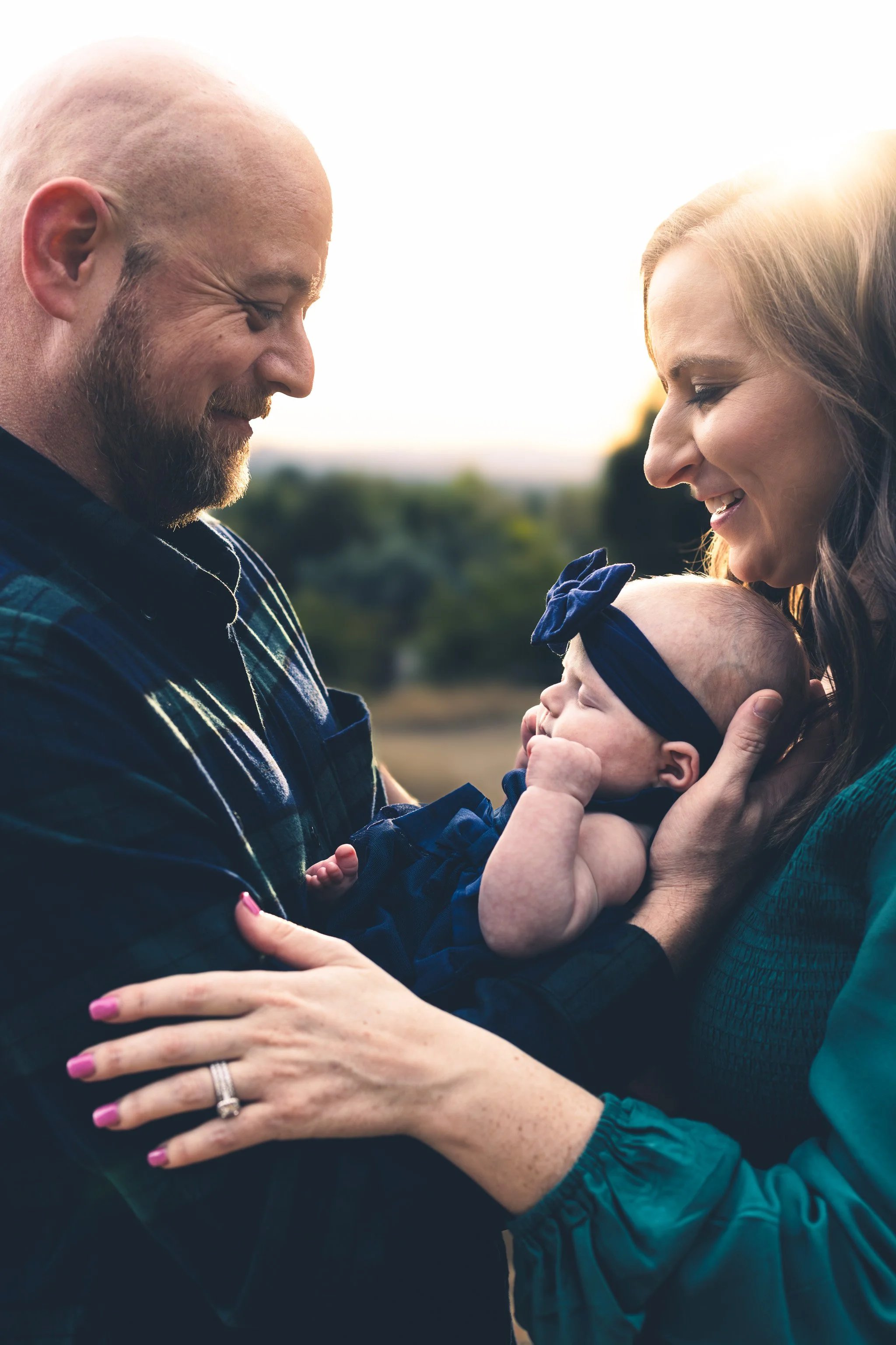 A family of three, a man, woman, and their baby girl, outdoors at sunset. The man is holding the baby, and the woman is gently touching the baby's hand. They are all smiling lovingly.