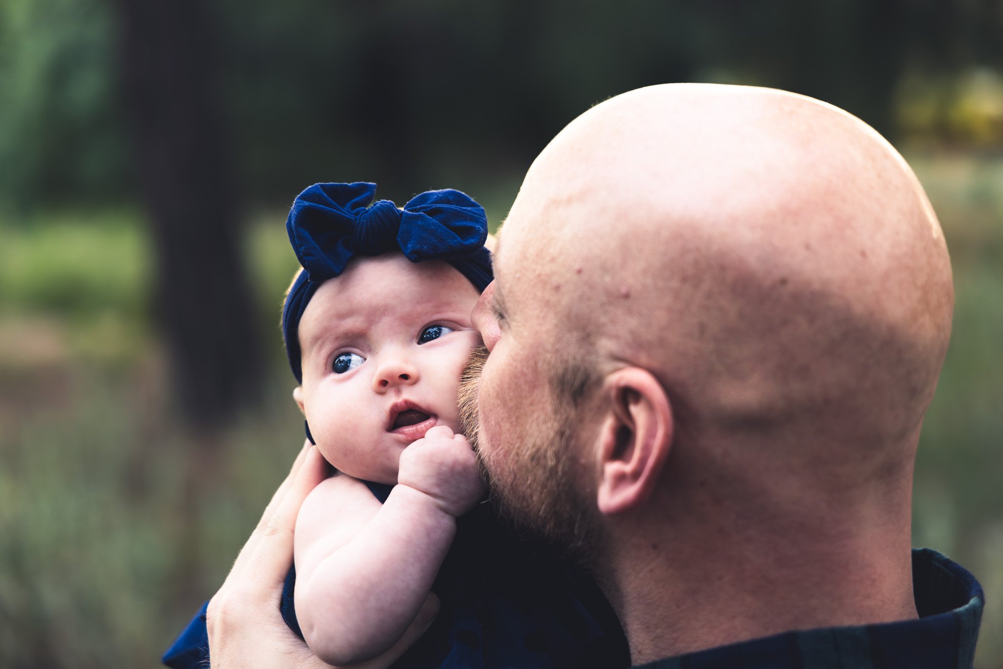 A man holding a baby girl outdoors, close-up with trees in the background. The baby has blue eyes, a dark blue headband with a bow, and is looking slightly upward. The man has a bald head and beard, and is kissing the baby on the cheek.