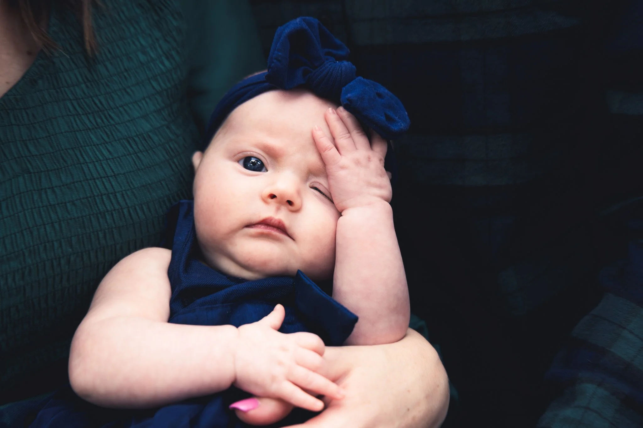 A baby girl with a blue headband and navy dress rests on a person's arm, touching her forehead with one hand and looking at the camera.