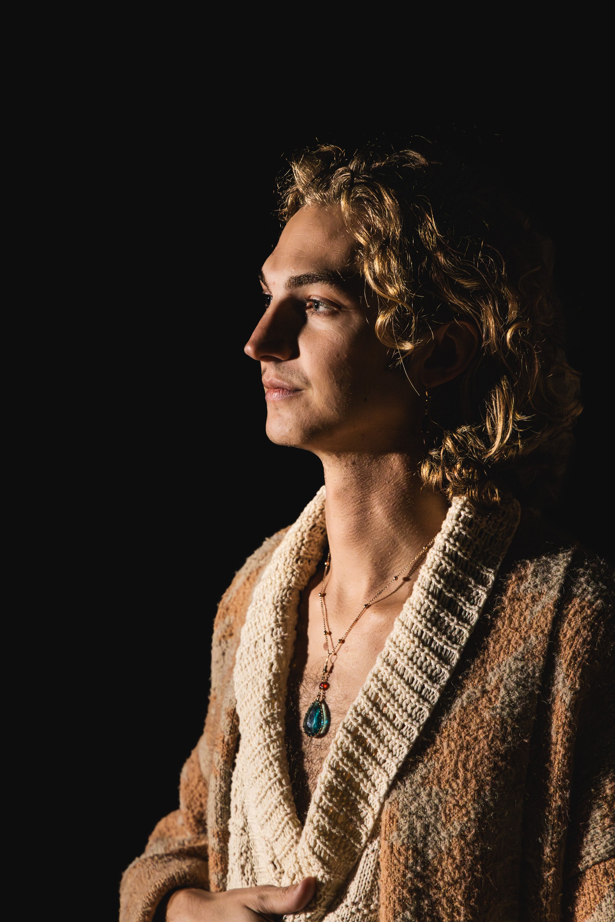 A young man with curly blond hair, wearing a beige knit sweater and a necklace with a large teardrop-shaped pendant, is looking to the left against a black background.