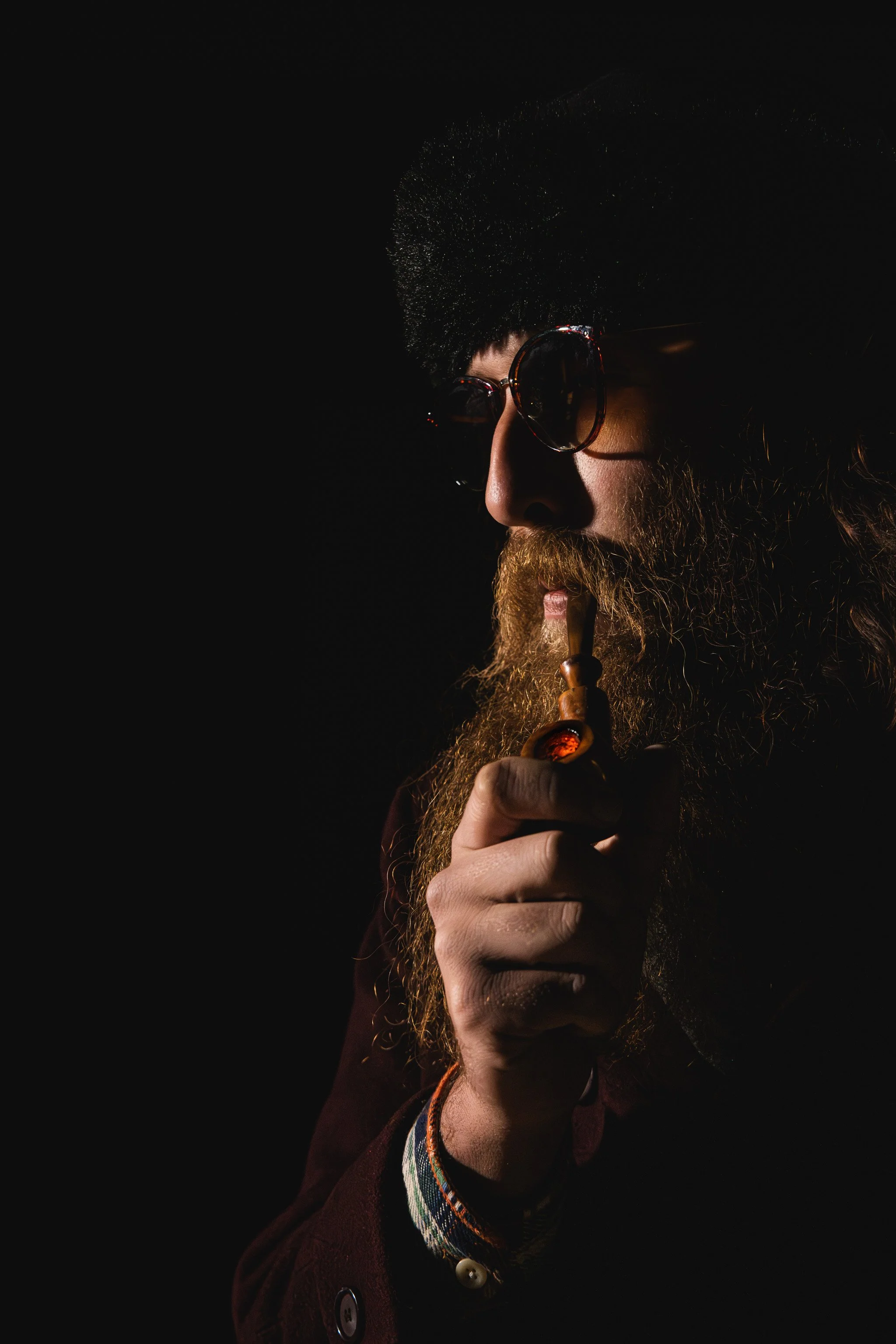 A man with a long beard, large sunglasses, and a furry hat smokes a pipe in a dark setting.