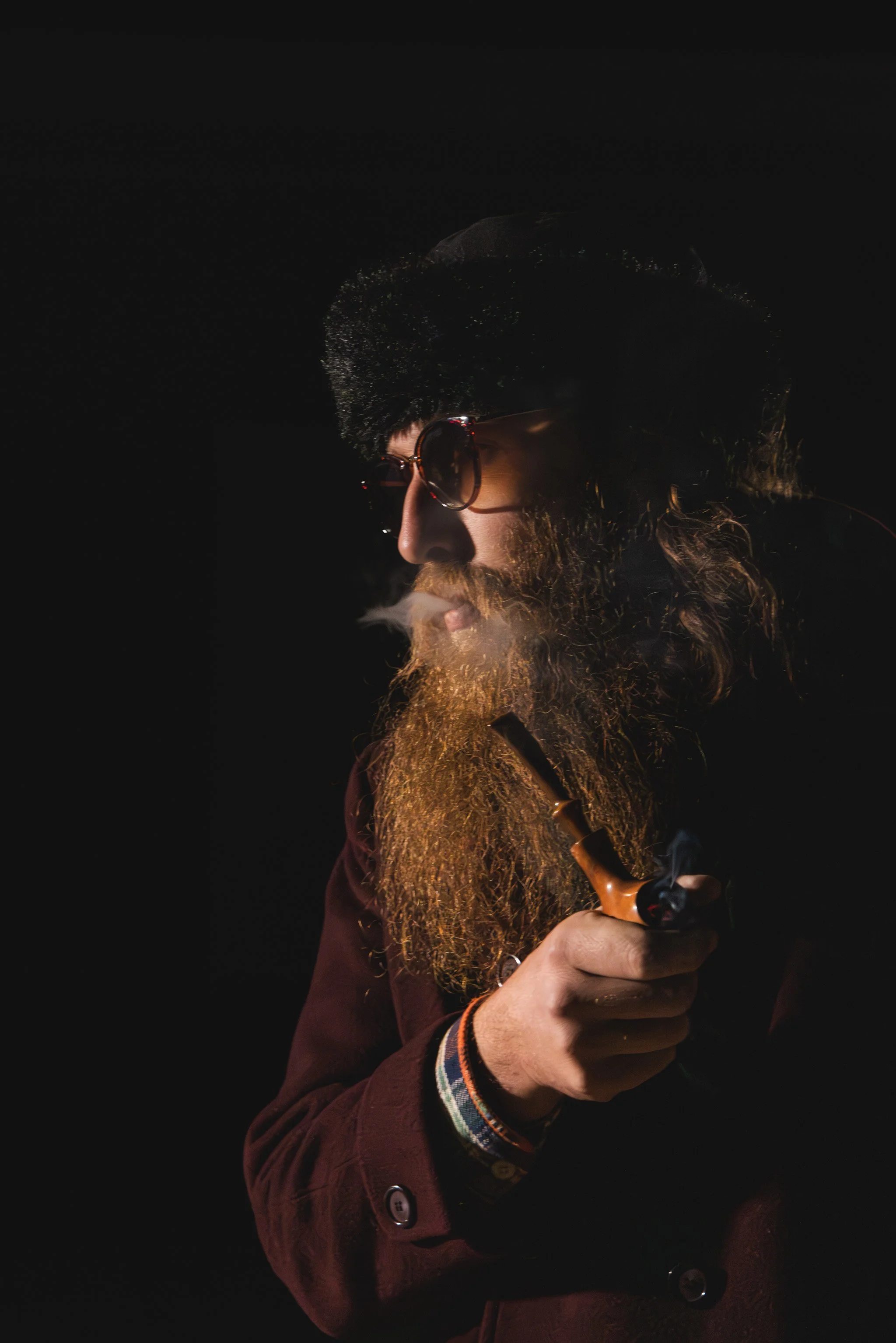 A man with a beard, sunglasses, and a fur hat smokes a pipe in a dark setting, with smoke visible.
