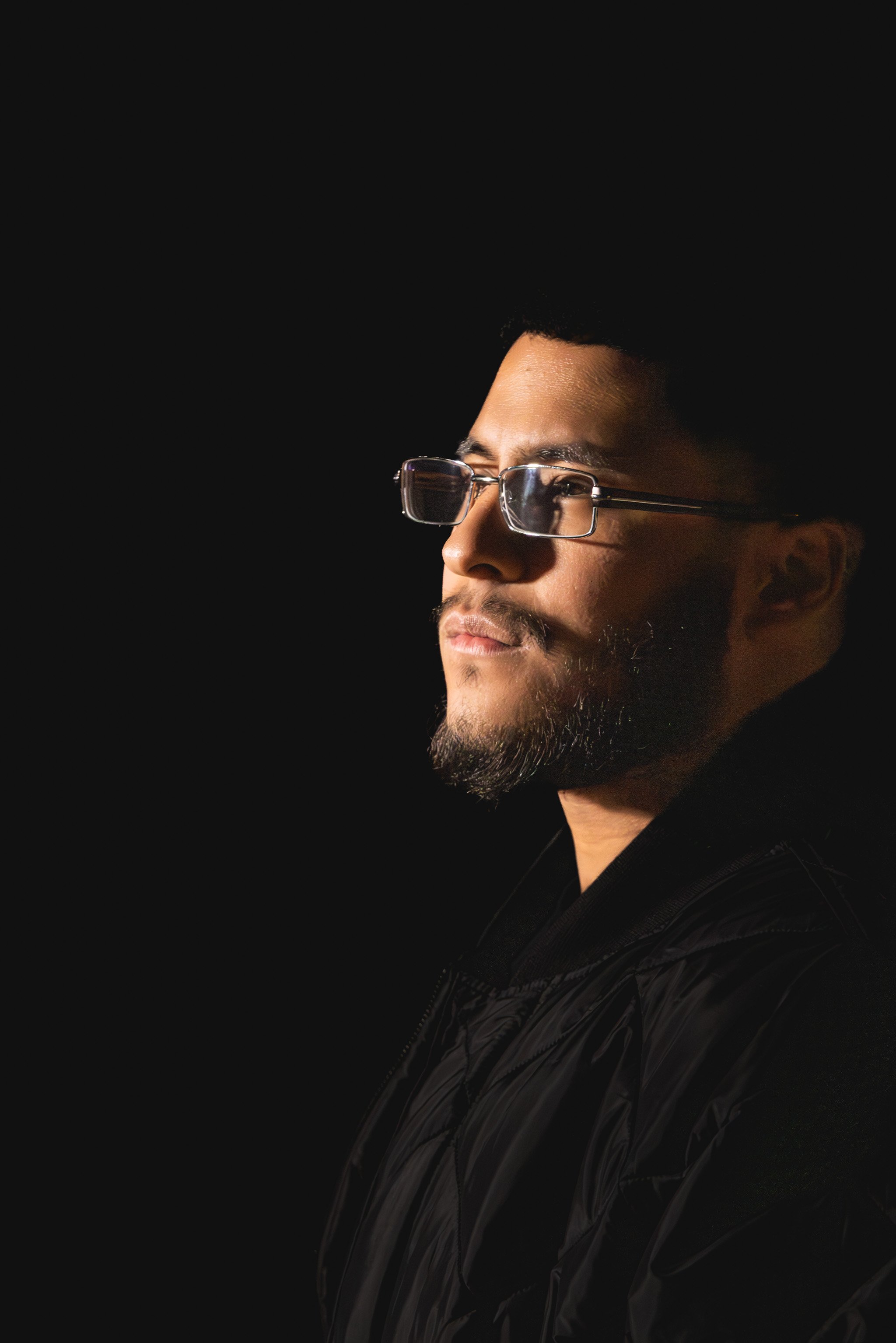 Side profile of a man with glasses, short black hair, and a beard, looking to the right against a black background.