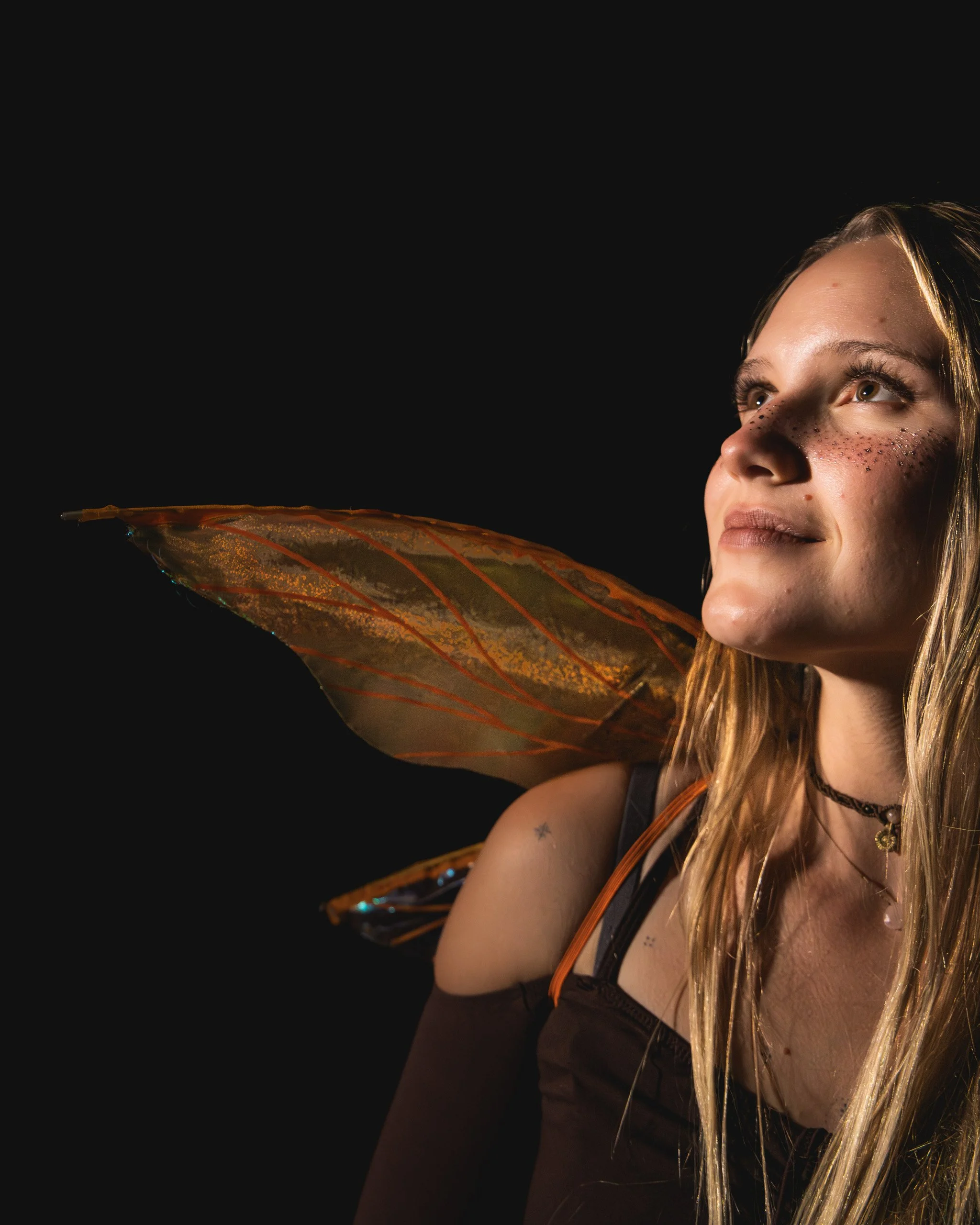 A woman with fairy wings, long blonde hair, and freckles, gazing upward, against a black background.