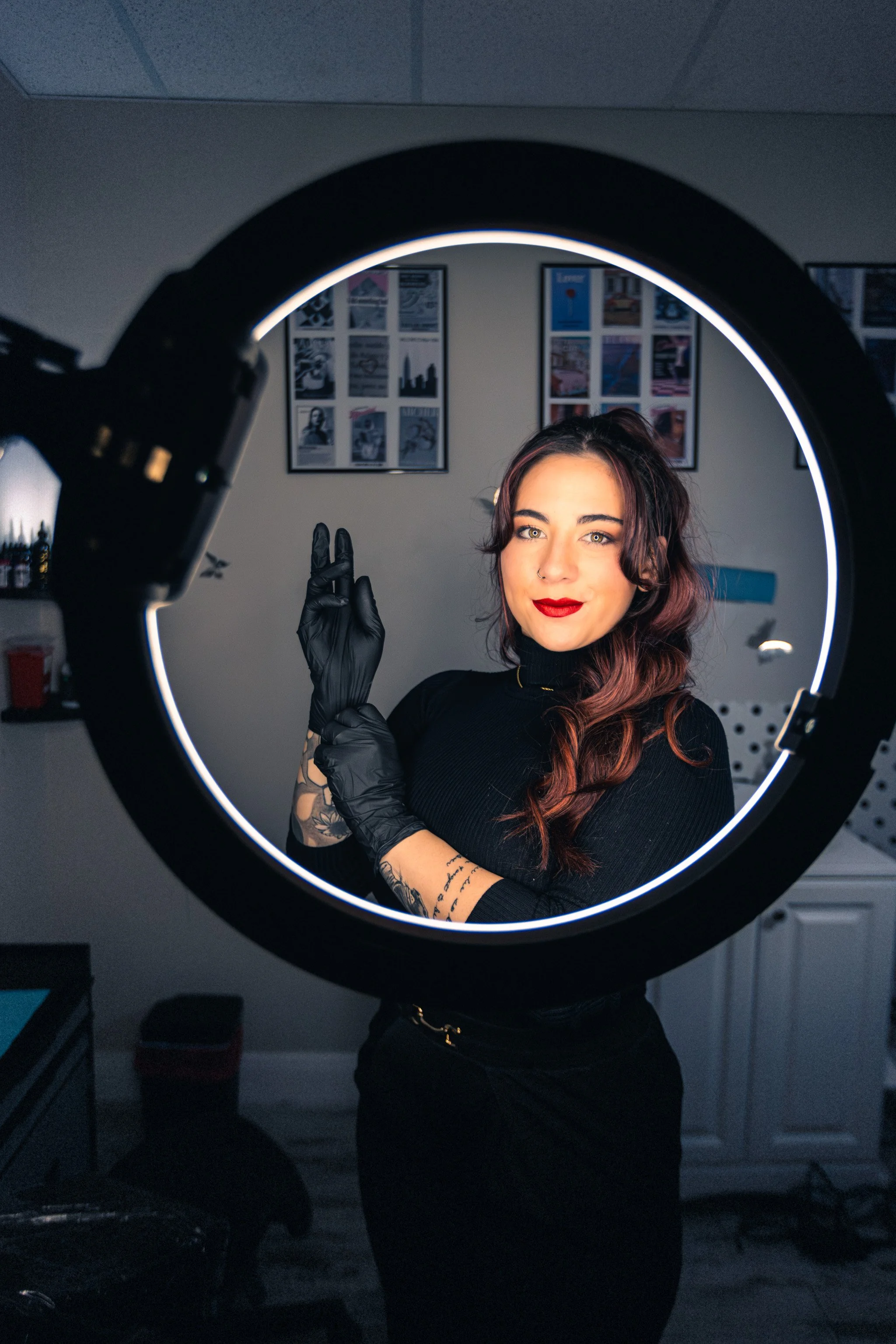A woman with long, wavy hair and red lipstick posing in front of a ring light, making a peace sign with her gloved hand in a room with framed pictures on the wall.
