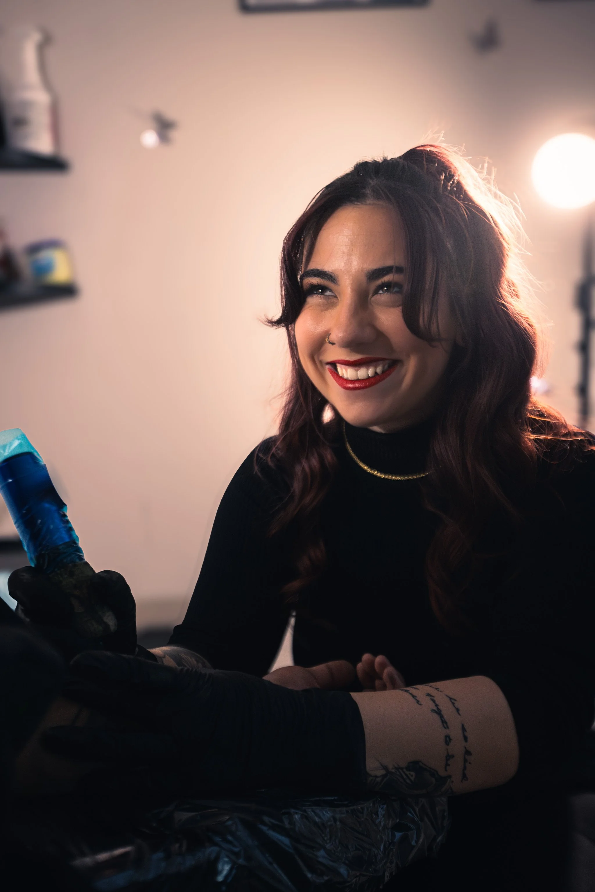 A woman with wavy brown hair, smiling, getting a tattoo on her forearm in a tattoo studio. She has a tattoo on her arm, is wearing a black shirt, red lipstick, and gold necklace, and is being attended by a tattoo artist in black gloves.
