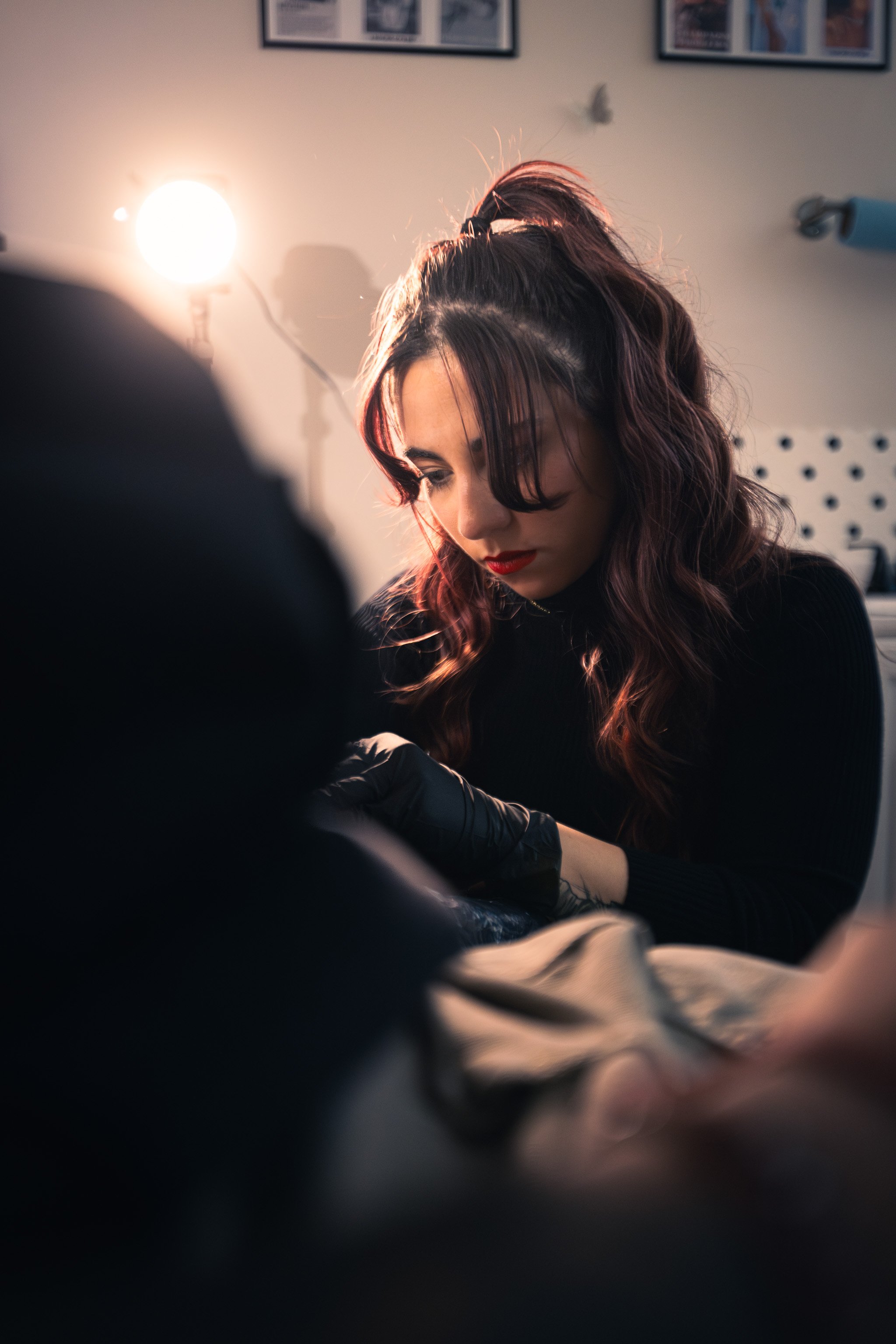 A woman with dark, wavy hair and red lipstick getting a tattoo from an artist wearing black gloves in a studio with framed photos on the wall and bright lighting.