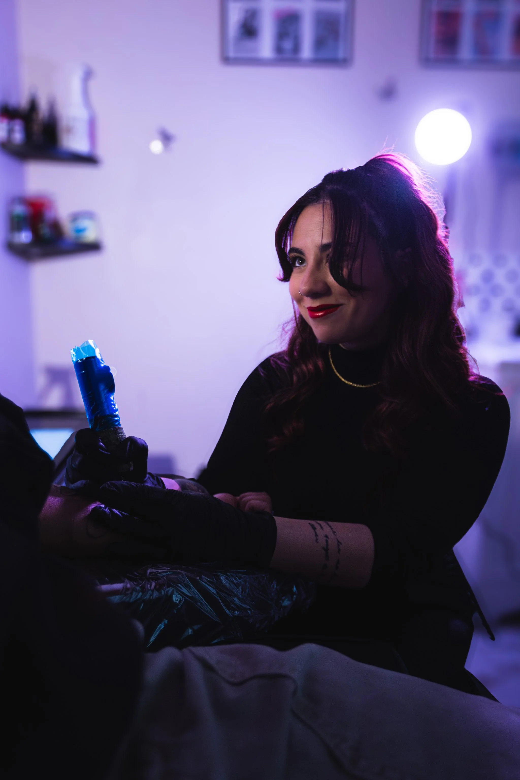 A woman with dark hair and red lipstick looks to the side while receiving a tattoo or piercing from someone wearing black gloves in a dimly lit room with purple and blue lighting.
