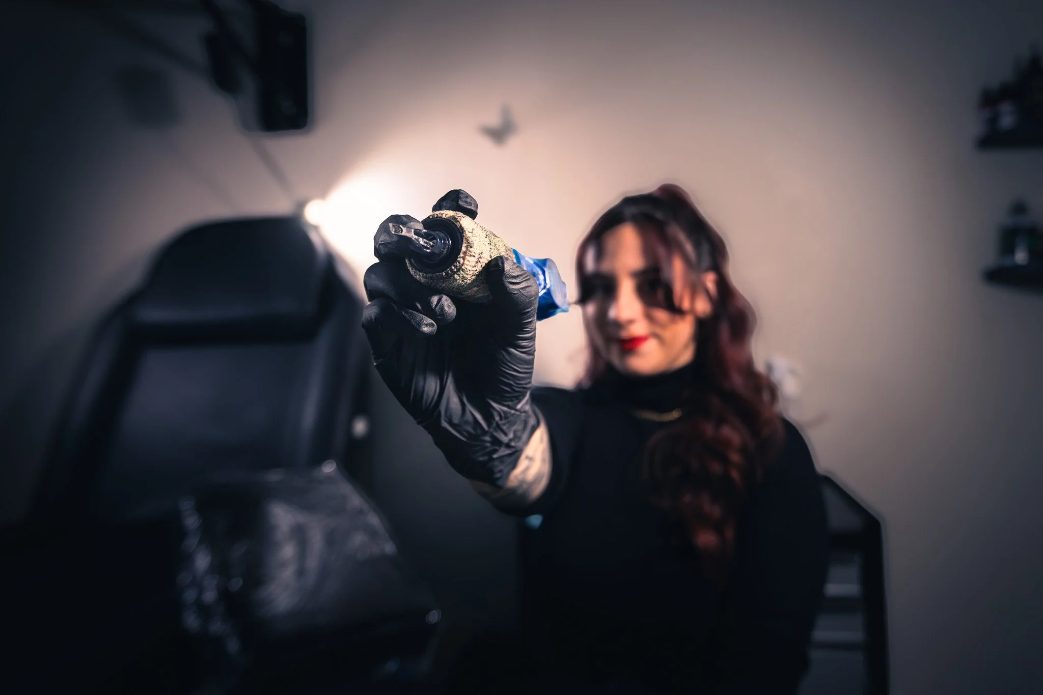 A woman with dark hair and red lipstick wearing a black long-sleeve shirt and black gloves holding a tattoo gun.