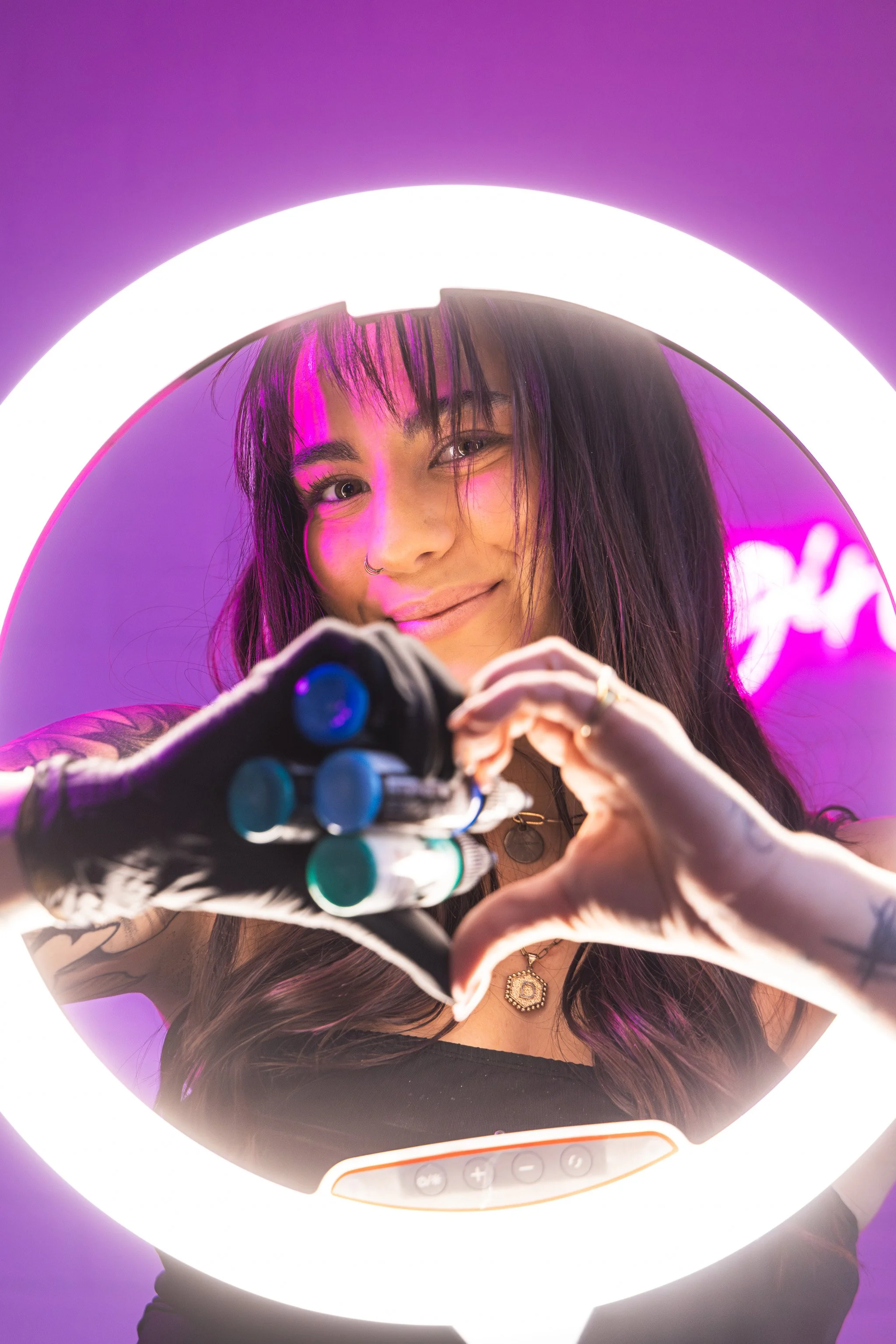 A young woman with dark hair, tattoos, and a nose piercing, smiling and making a heart shape with her fingers through a circular mirror with built-in LED lights. The background has pink and purple neon lights.