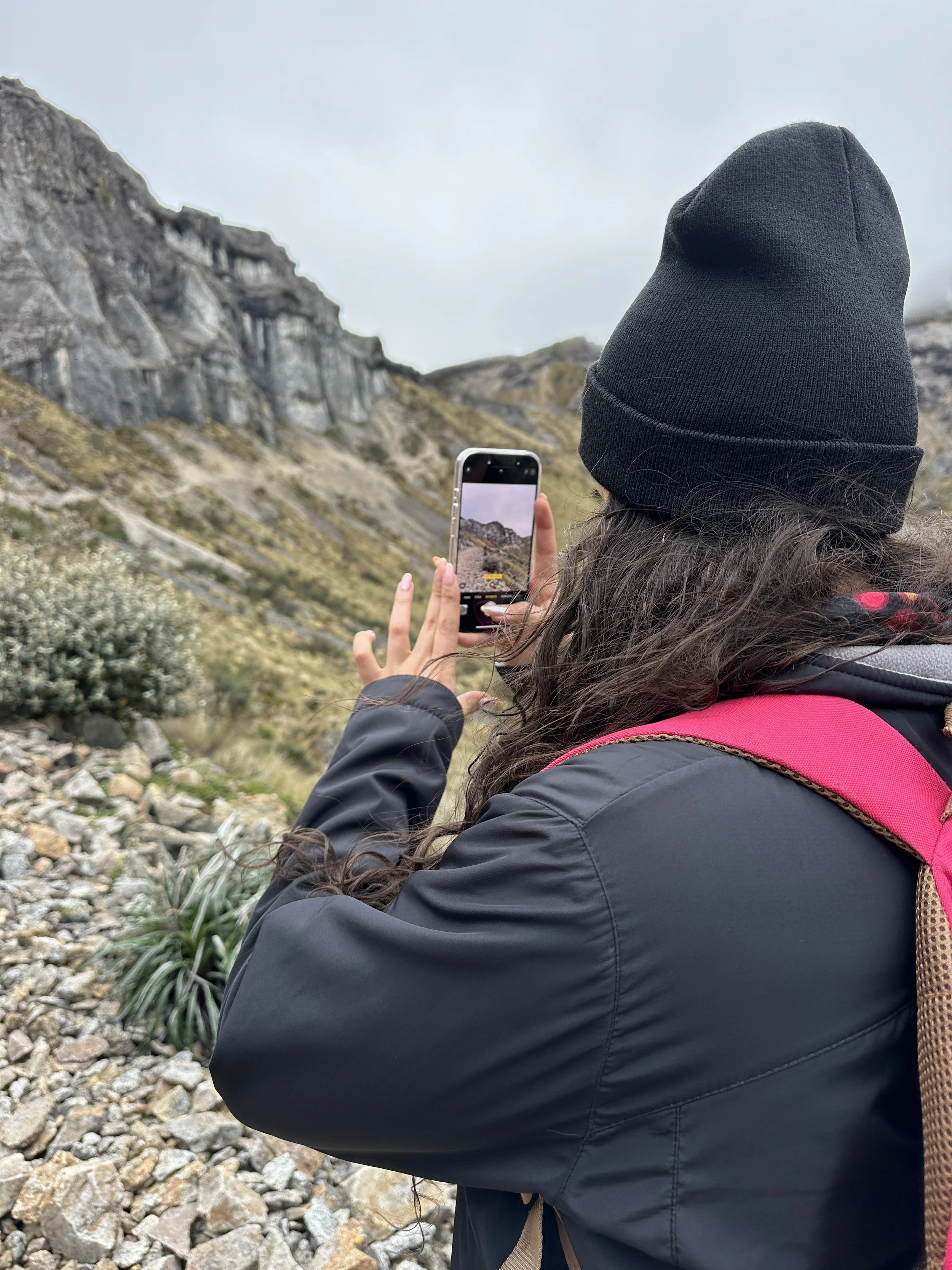 Una mujer con gorro negro y chaqueta oscura toma una foto con su teléfono móvil en un paisaje montañoso con rocas y vegetación, lleva una mochila rosa.