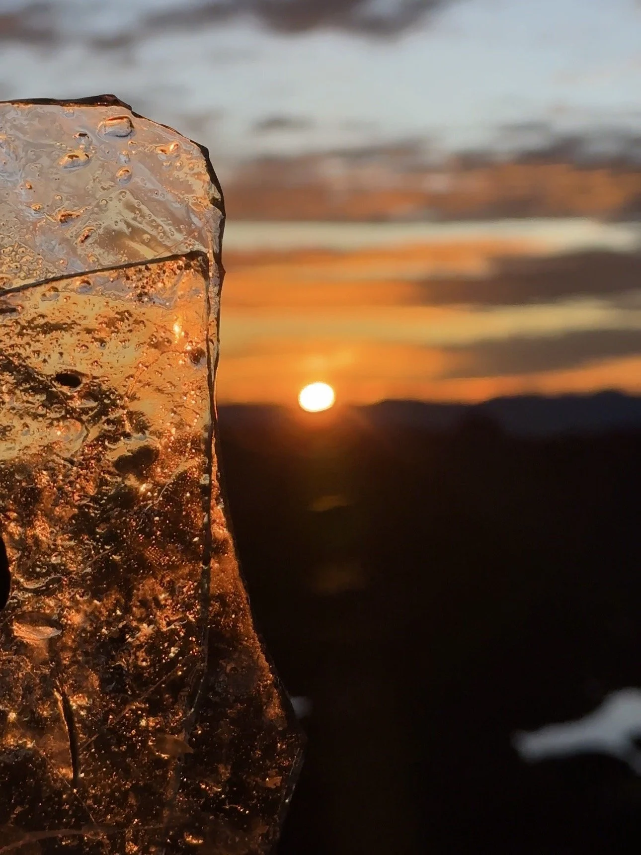 Un trozo de hielo transparente en primer plano, reflejando el atardecer con el sol en el horizonte, en un paisaje de montaña.