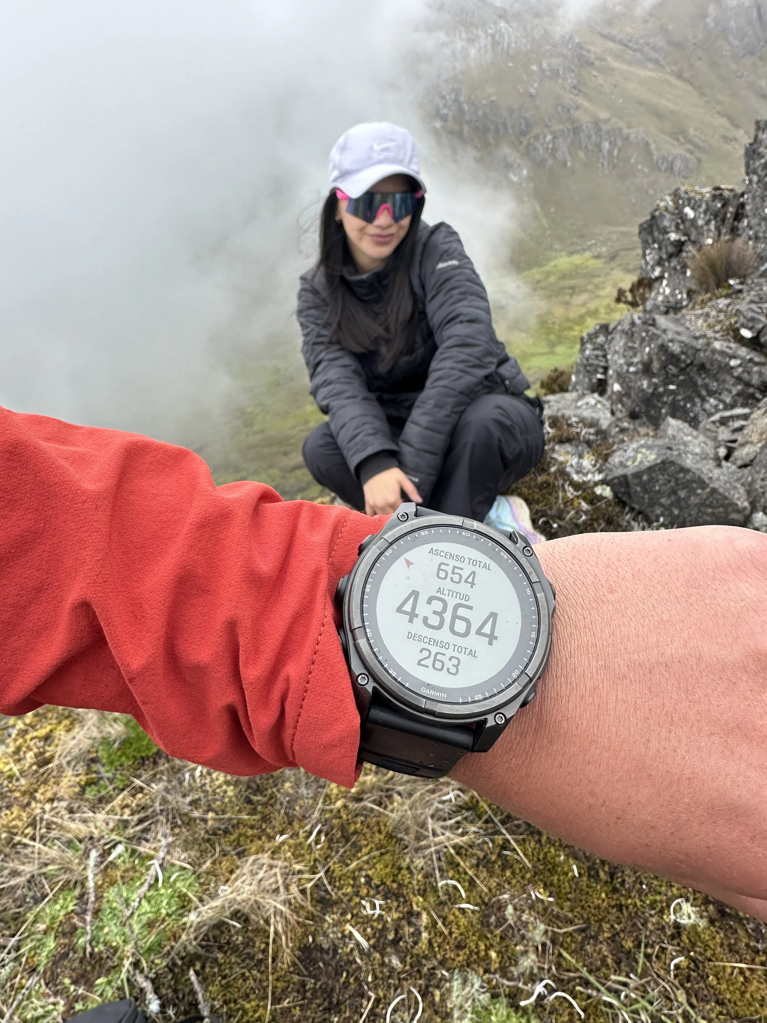 Mujer en ropa de senderismo en una zona montañosa con neblina, mostrando un reloj GPS que indica una altitud de 4364 metros y un ascenso total de 654 metros.
