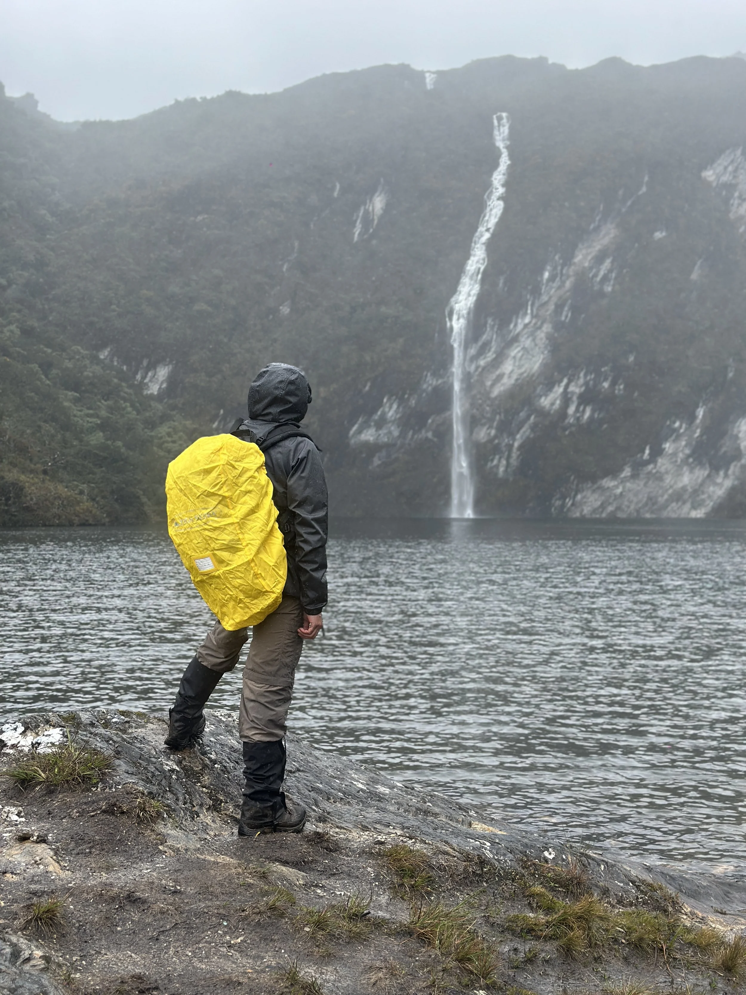 Persona con ropa de lluvia y mochila amarilla observa una cascada desde la orilla de un lago rodeado de montañas.
