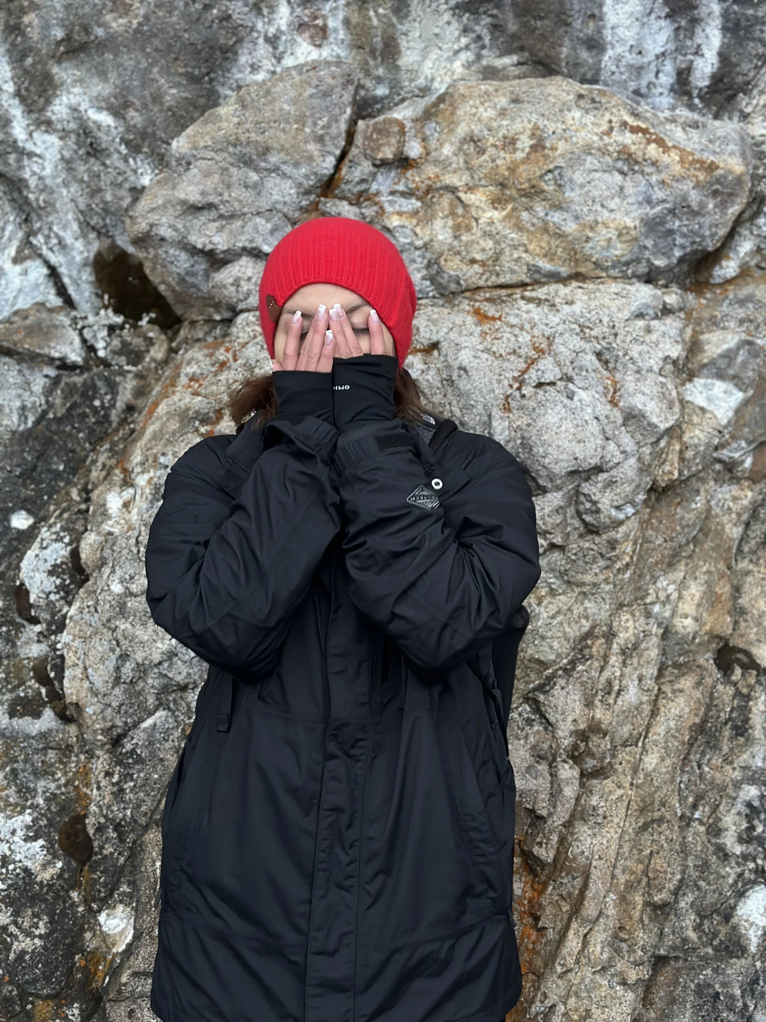 Mujer con gorro rojo y chaqueta negra cubriéndose la cara con las manos frente a una roca grande.