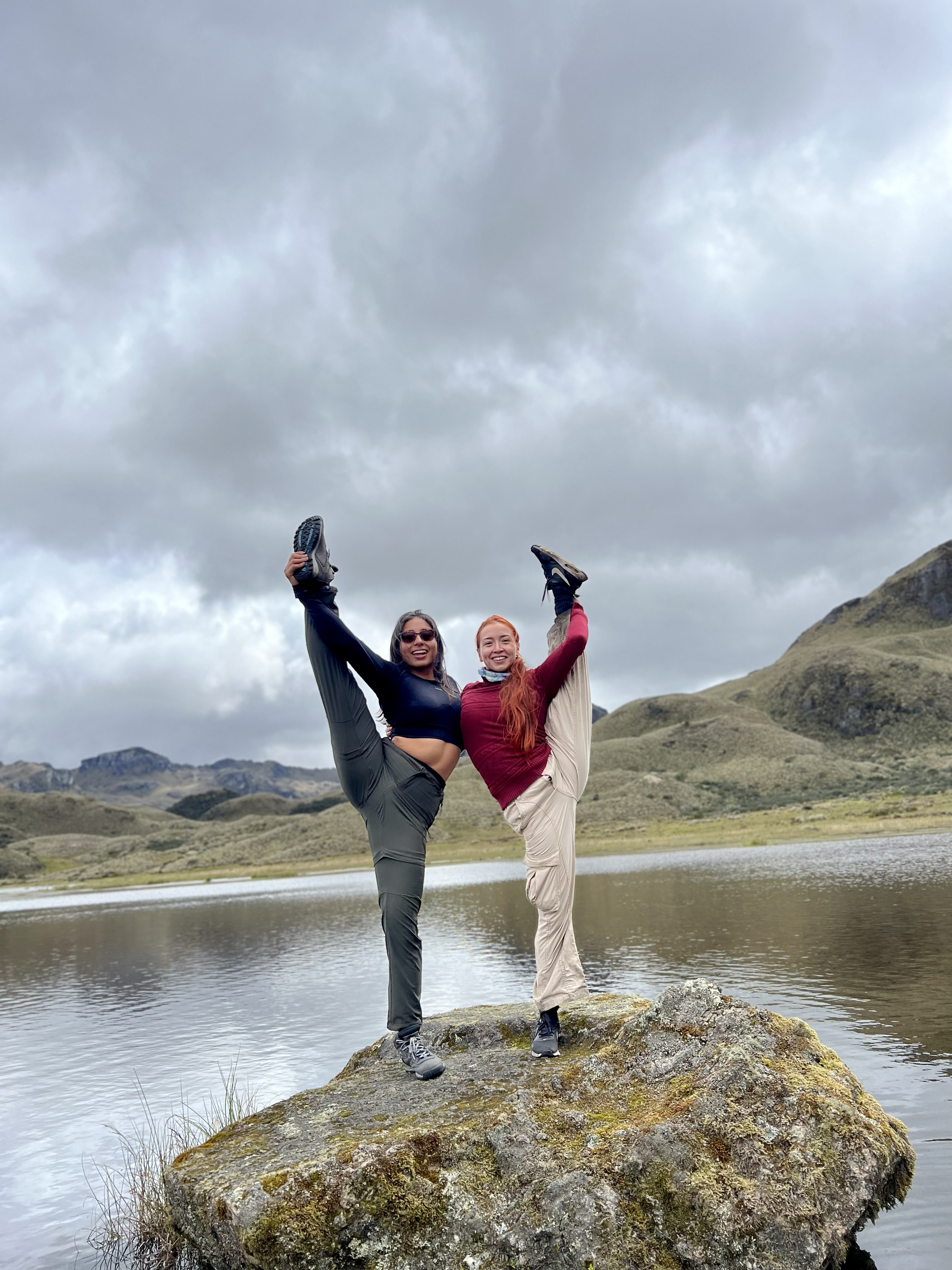 Dos mujeres con ropa deportiva haciendo un ejercicio de estiramiento en equilibrio sobre una roca en un lago rodeado de montañas y cielos nublados.
