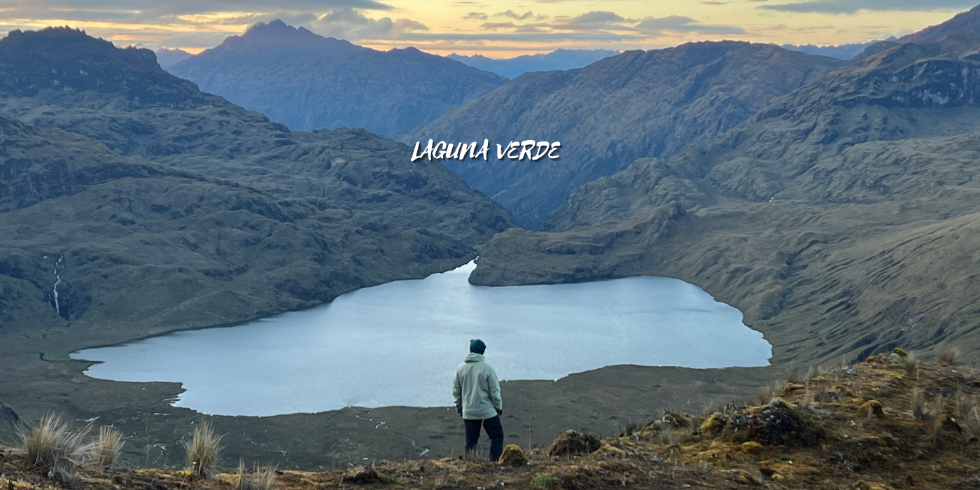 Persona observando laguna verde en un paisaje montañoso al atardecer con nubes en el cielo