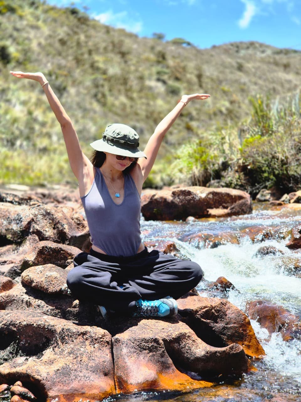 Mujer sentada cruzada en una roca junto a un río, haciendo yoga o meditación, con brazos extendidos hacia arriba, sombrero, gafas y ropa casual en un paisaje de montañas y cielo azul.