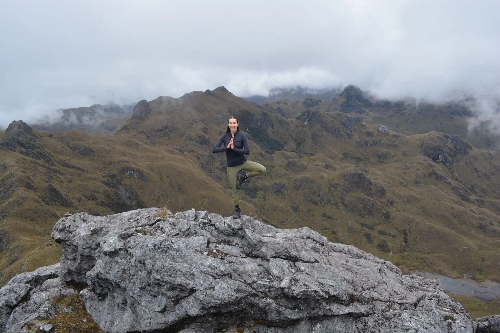 Mujer practicando yoga en un risco en una montaña con neblina y áreas verdes.