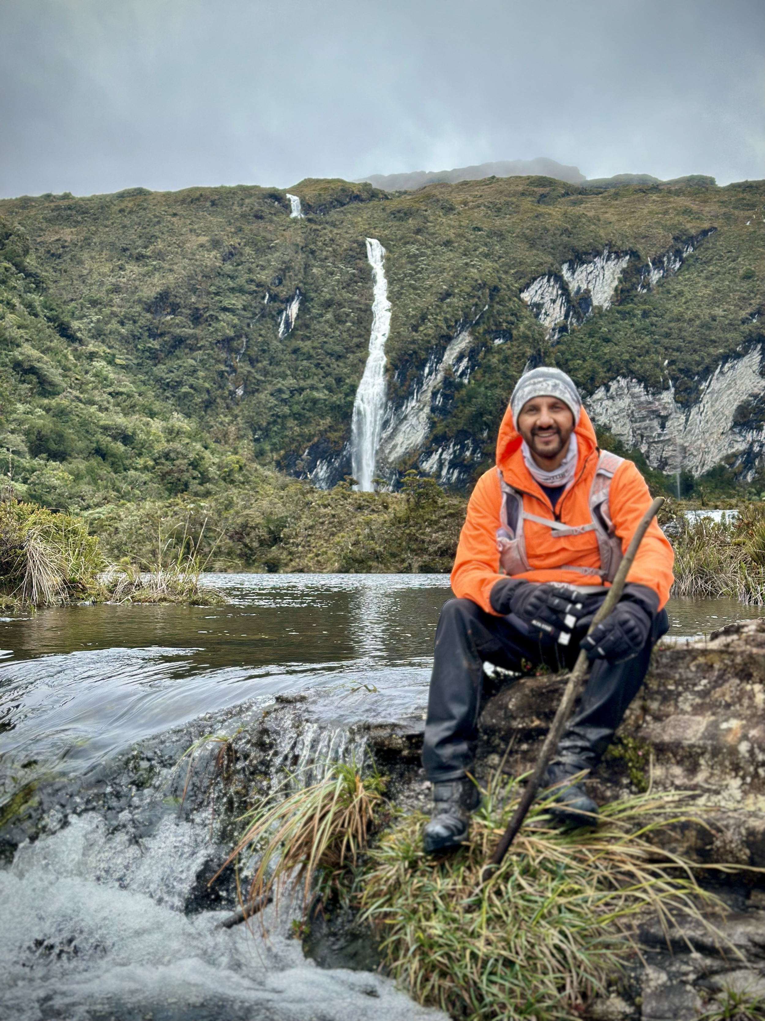 Hombre sonriente sentado sobre una roca junto a un río, con una cascada en la montaña de fondo, vistiendo ropa de senderismo naranja y gris.