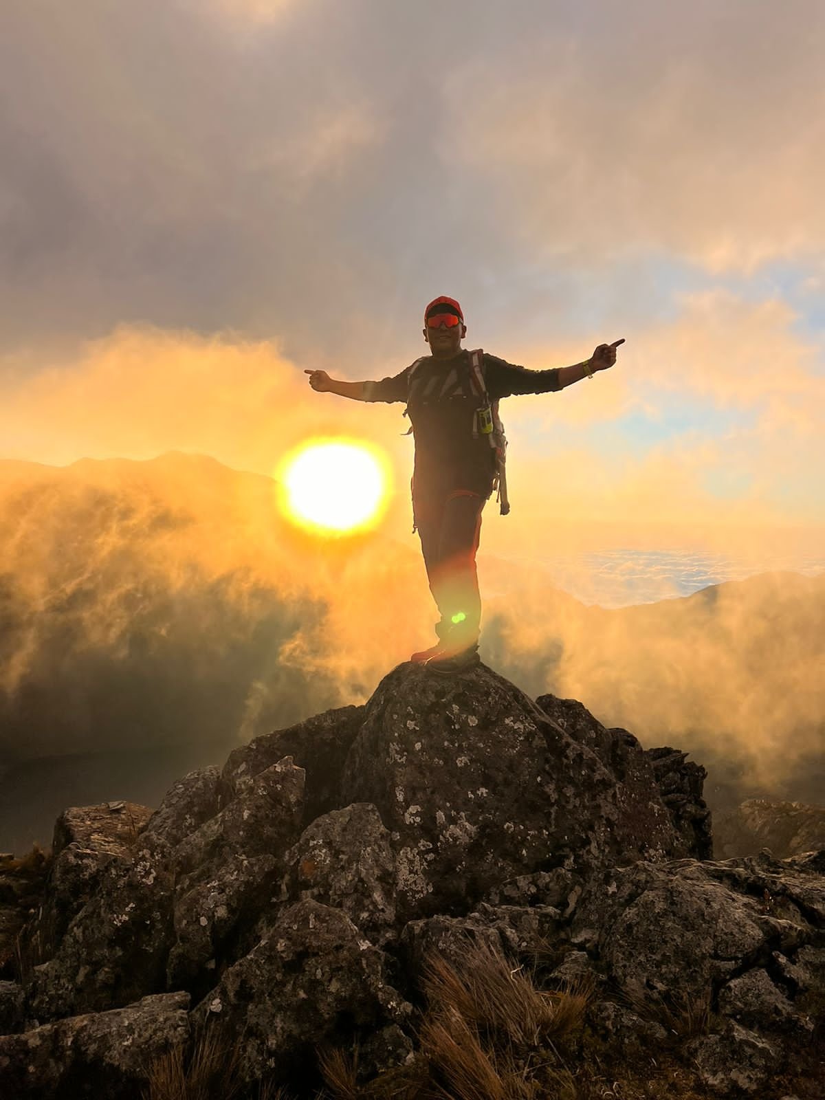 Persona haciendo senderismo en una montaña durante el atardecer, de pie sobre una roca con los brazos abiertos.