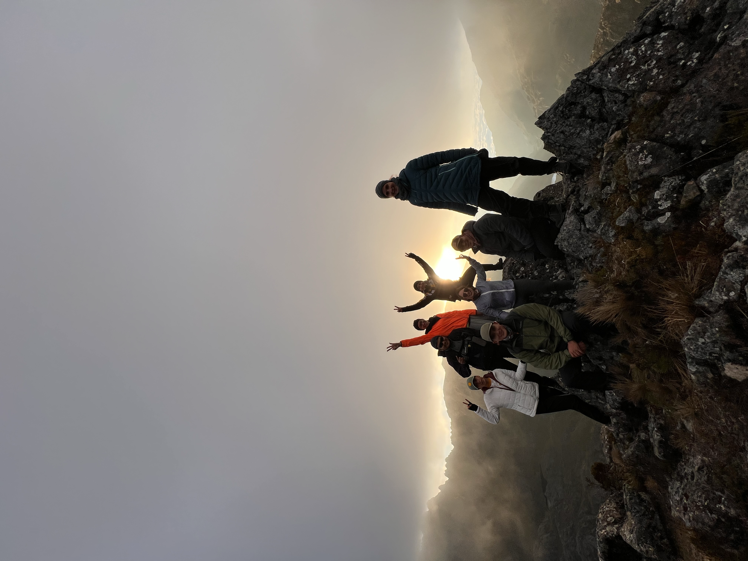 Grupo de personas en la cima de una montaña al atardecer, haciendo gestos de paz y celebración.
