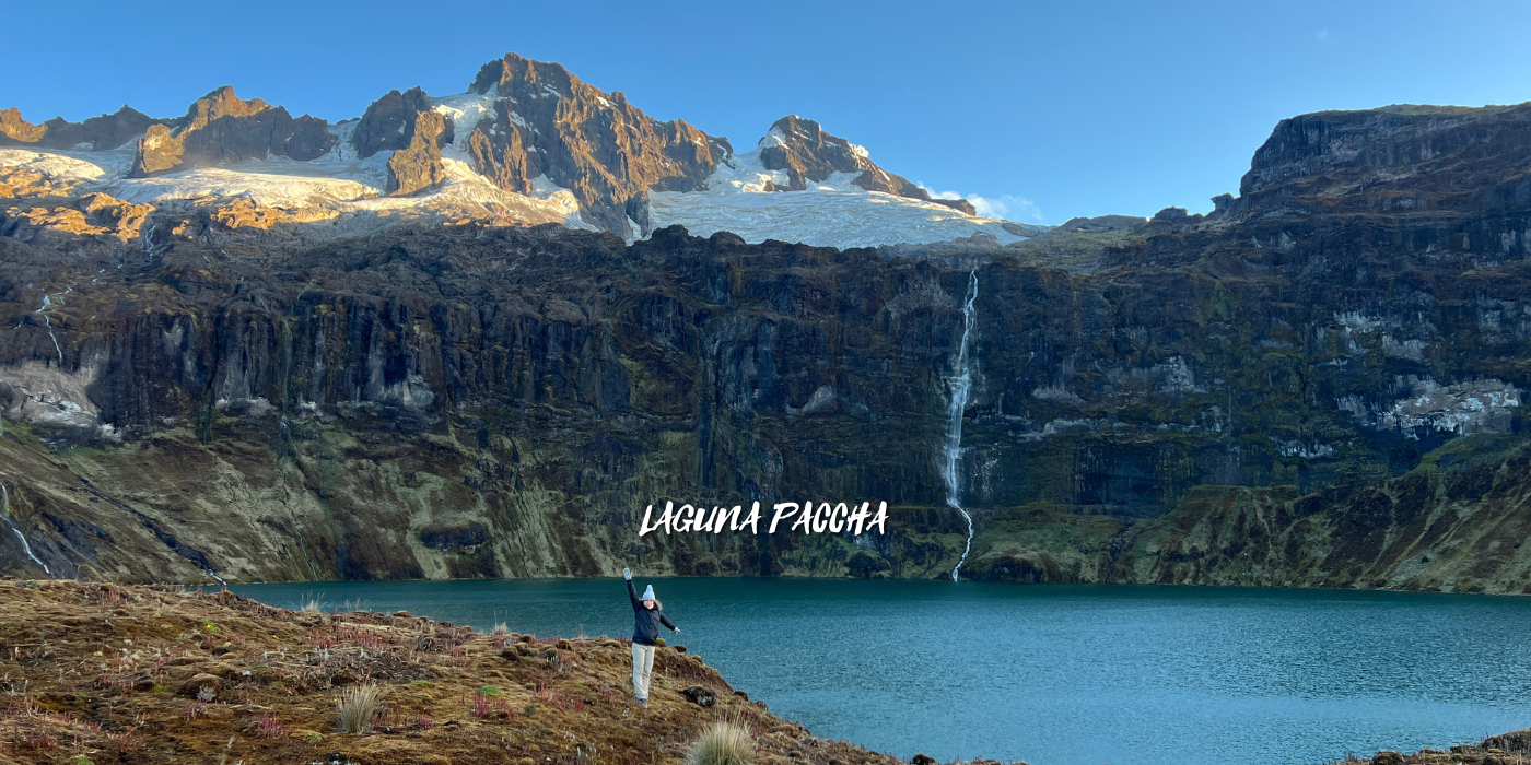 Persona con gorra y chaqueta en un paisaje montañoso frente a un lago, con cascadas en las montañas al fondo, en Laguna Pachcha.