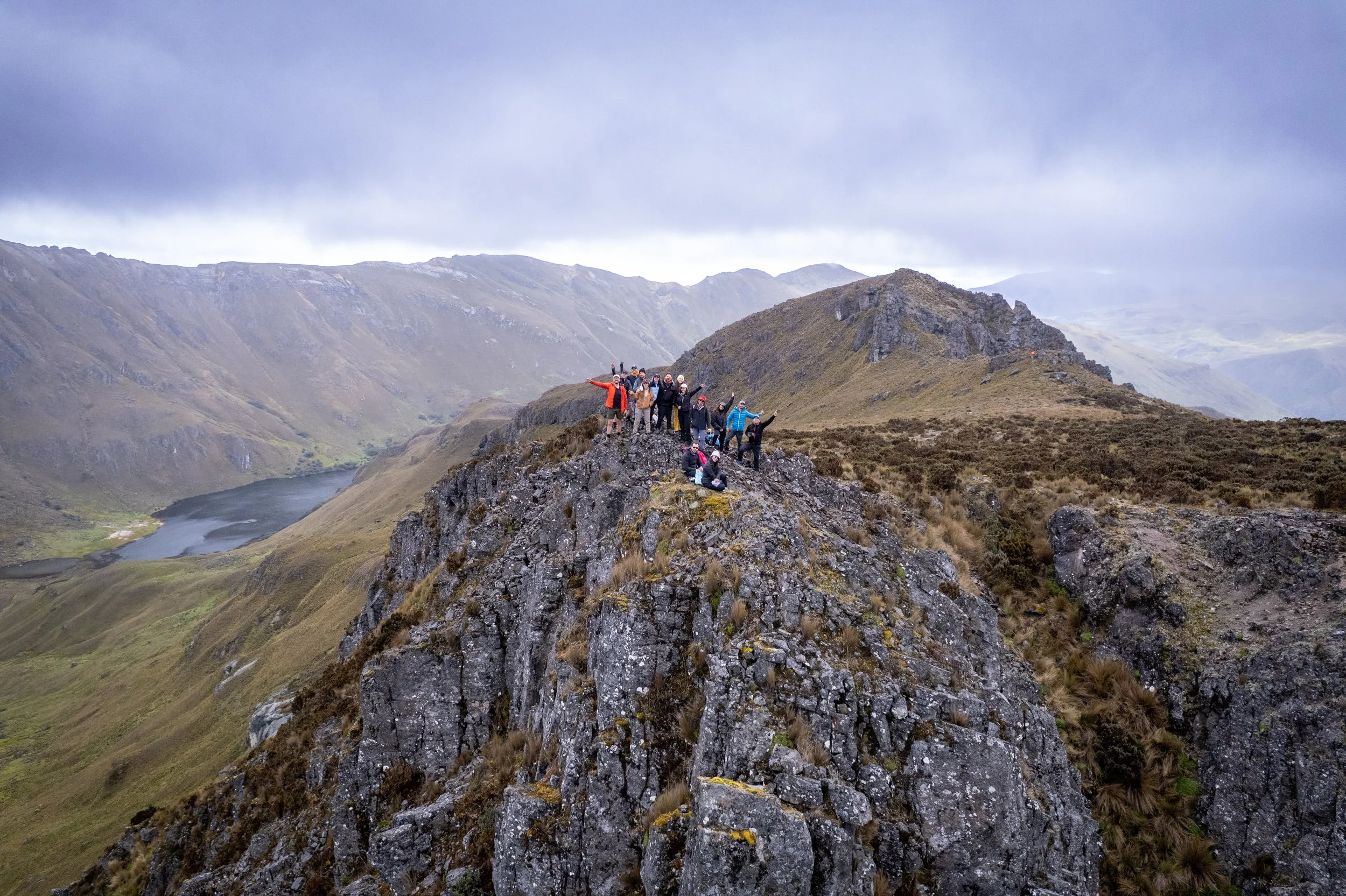 Grupo de personas en la cima de una formación rocosa en un paisaje montañoso con lagos y montañas en el fondo, nublado y con clima frío.