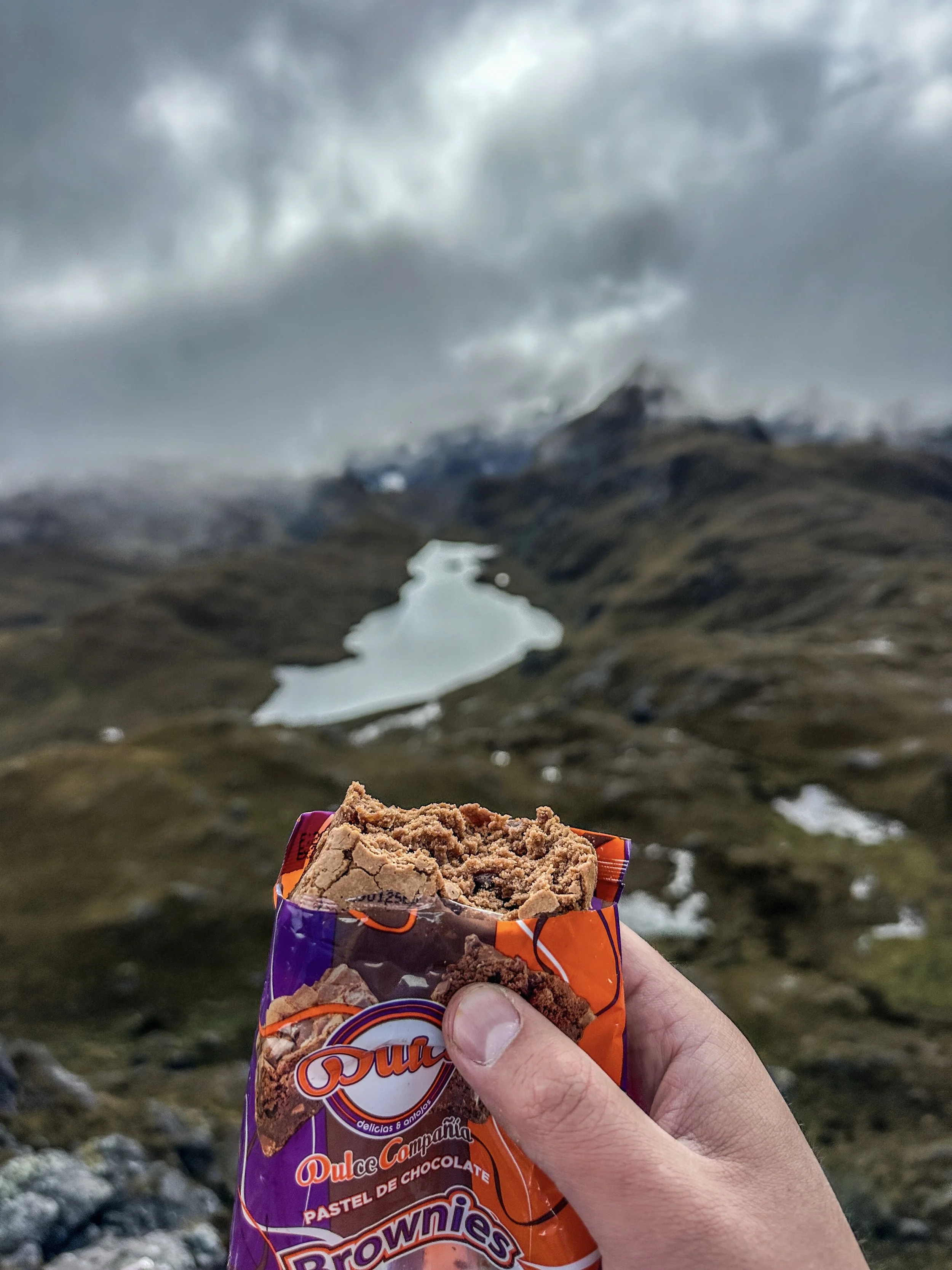 Una mano sostiene una bolsa de brownies de chocolate en un paisaje montañoso con un lago y nubes oscuras en el fondo.