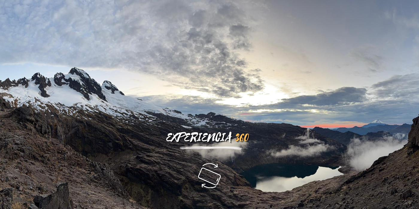 Paisaje montañoso con picos nevados, lago en medio, nubes y un cielo parcialmente despejado en un entorno frío y escarpado.
