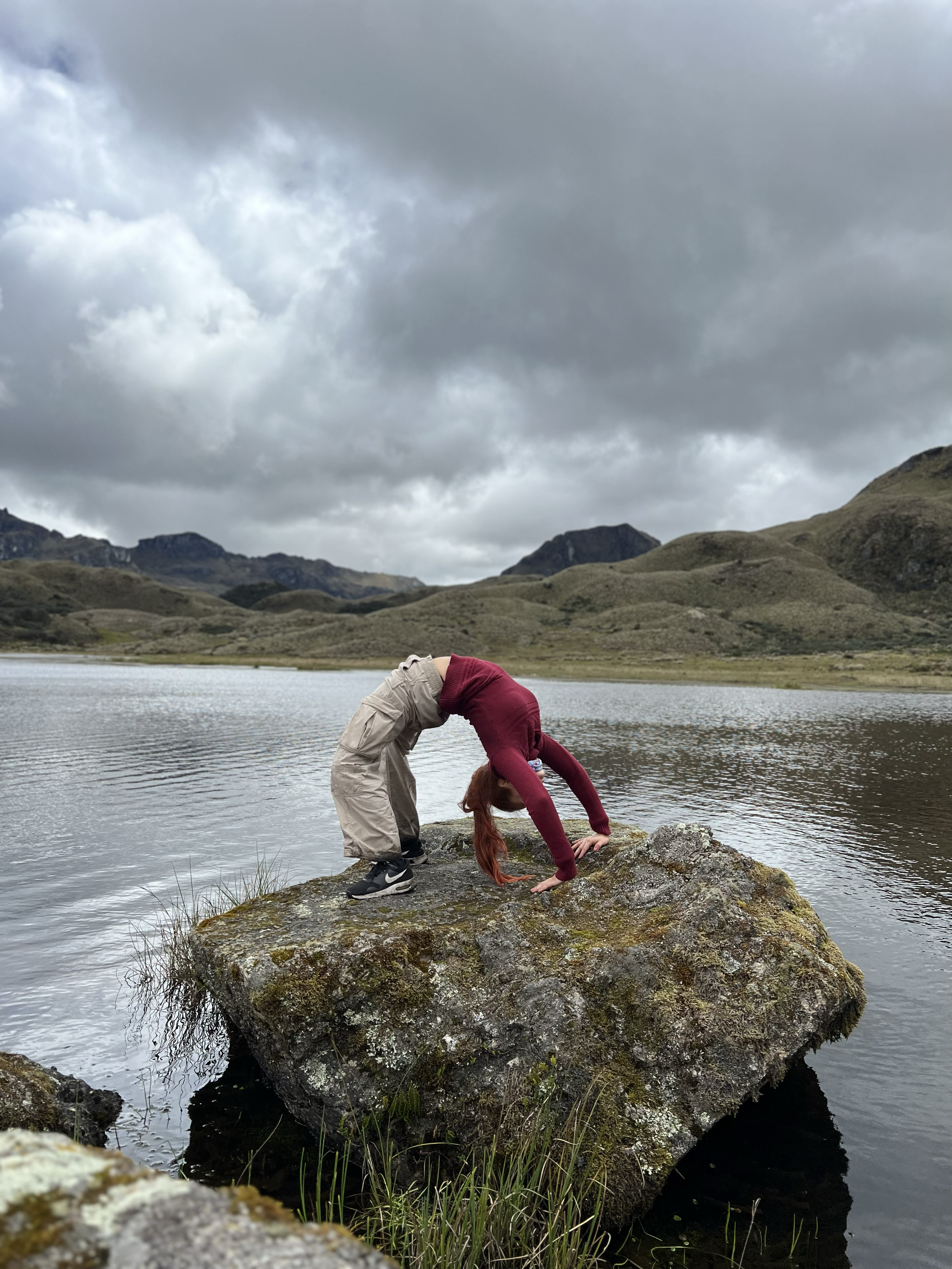 Persona con camiseta roja y pantalones beige doblando hacia atrás, haciendo una postura en una roca en el agua de un lago en un paisaje montañoso con nubes grises.