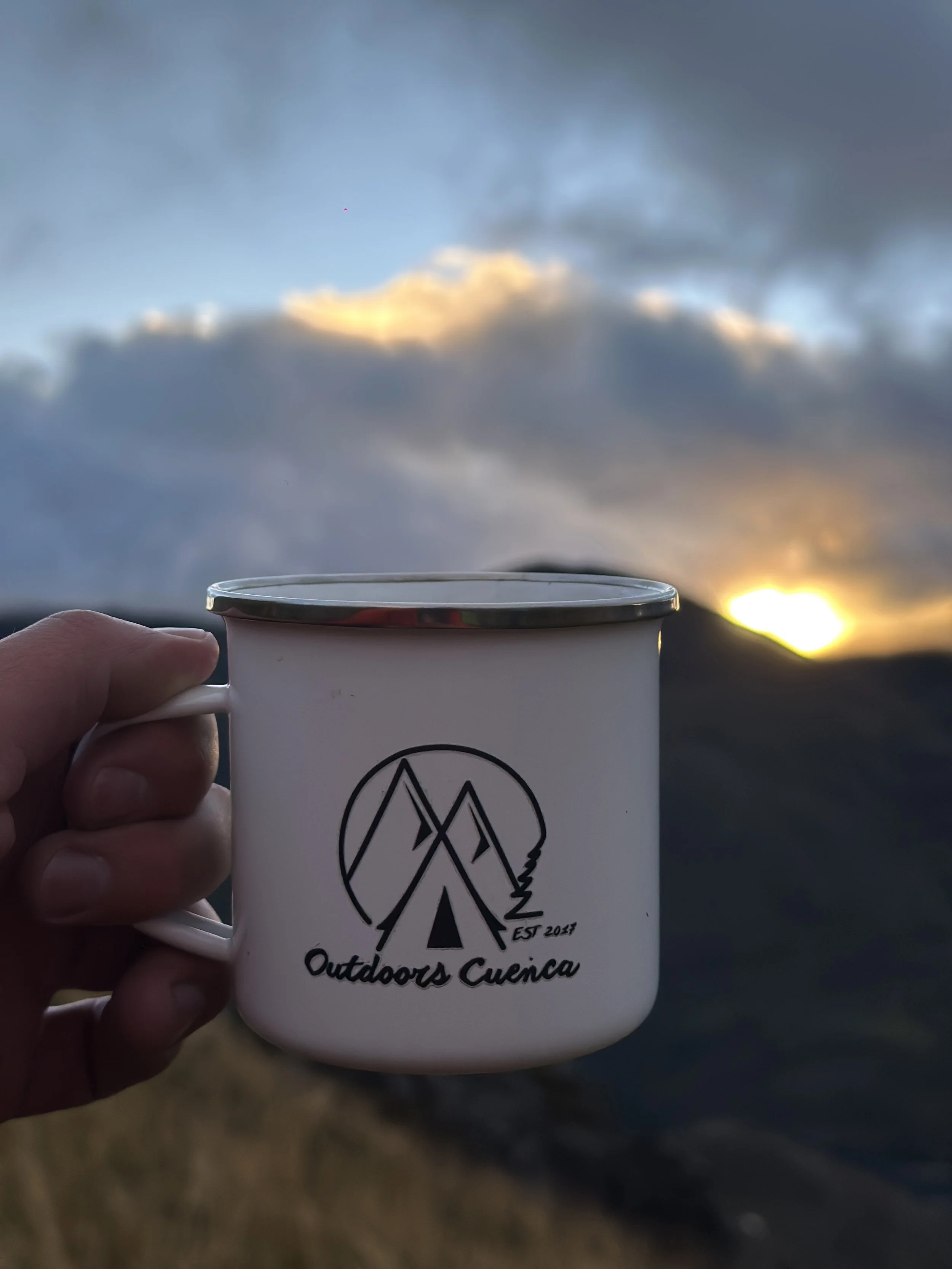 Taza de camping de color blanco con diseño de montañas, árbol y la leyenda 'Outdoors Cuenca' y 'EST 2021', sostenida por una mano con paisaje de montañas y cielo nublado al atardecer en el fondo.