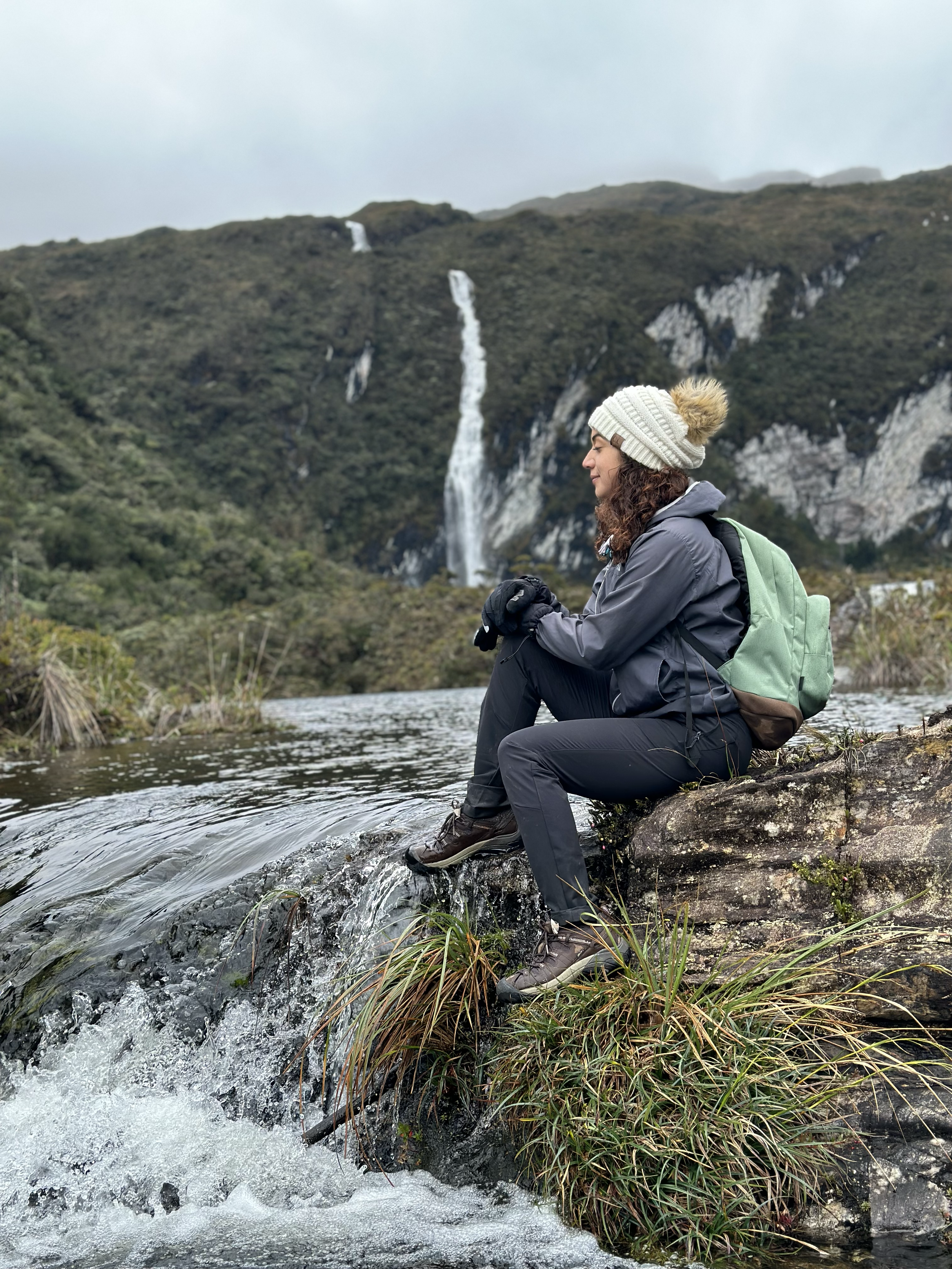 Mujer sentada en una roca cerca de un río con un paisaje de montañas y cascadas en el fondo.