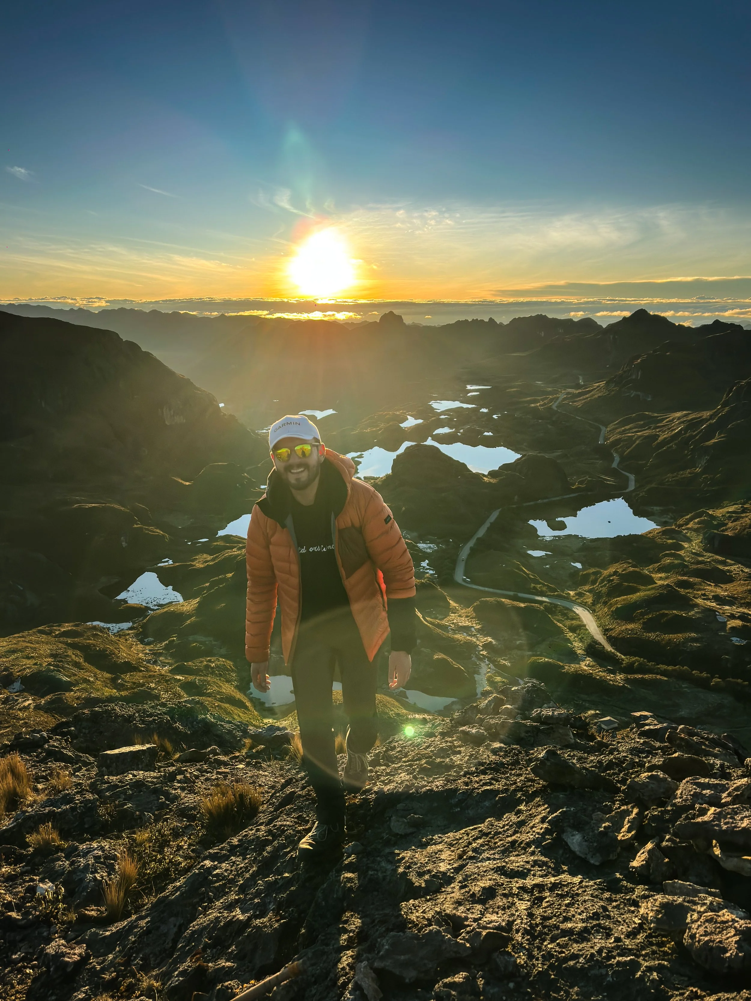 Un hombre sonriendo con gafas de sol y chaqueta naranja, escalando una montaña con lagunas y un valle extenso al atardecer.