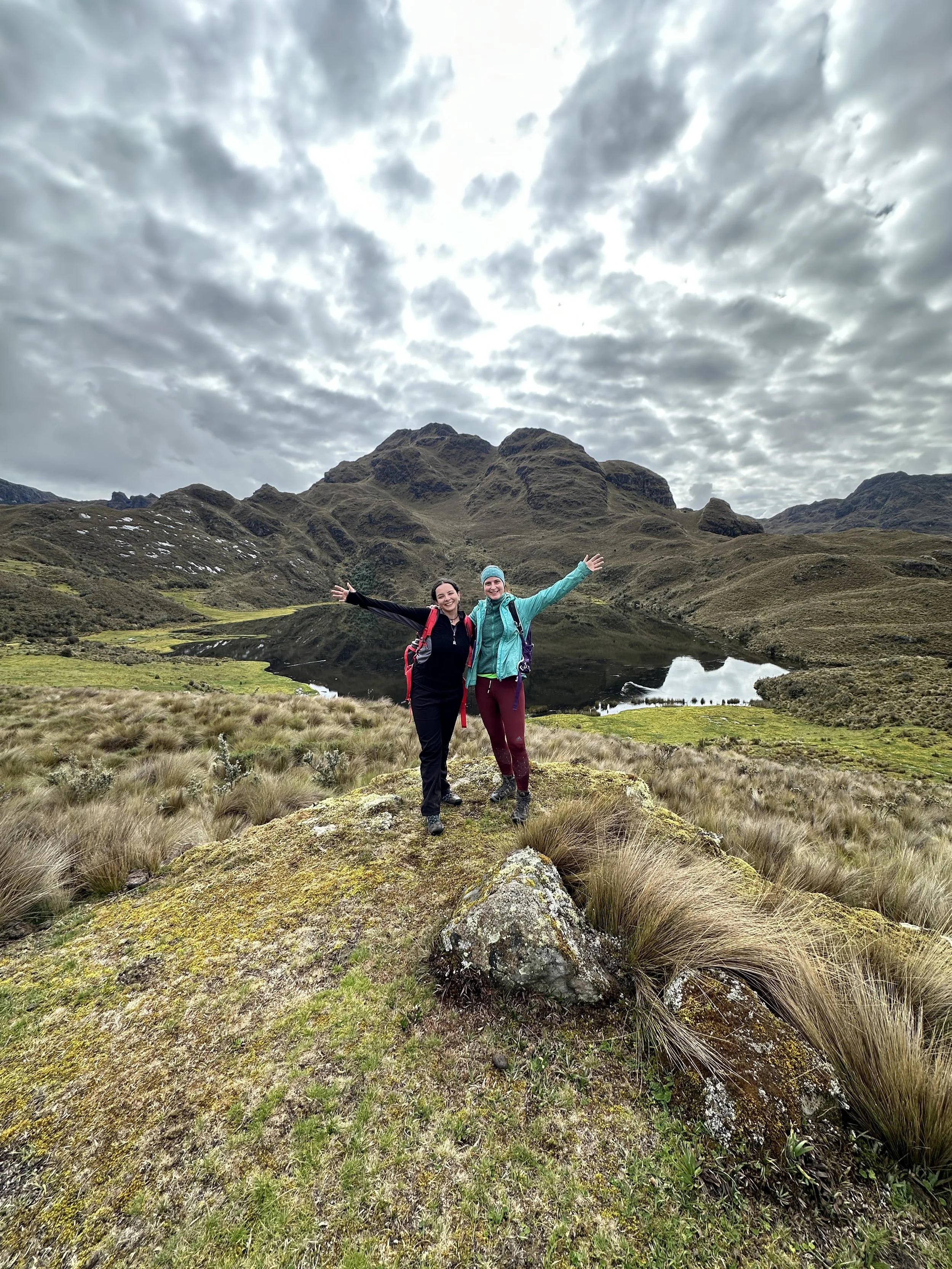 Dos personas de pie en un paisaje montañoso con un lago y nubes en el cielo.