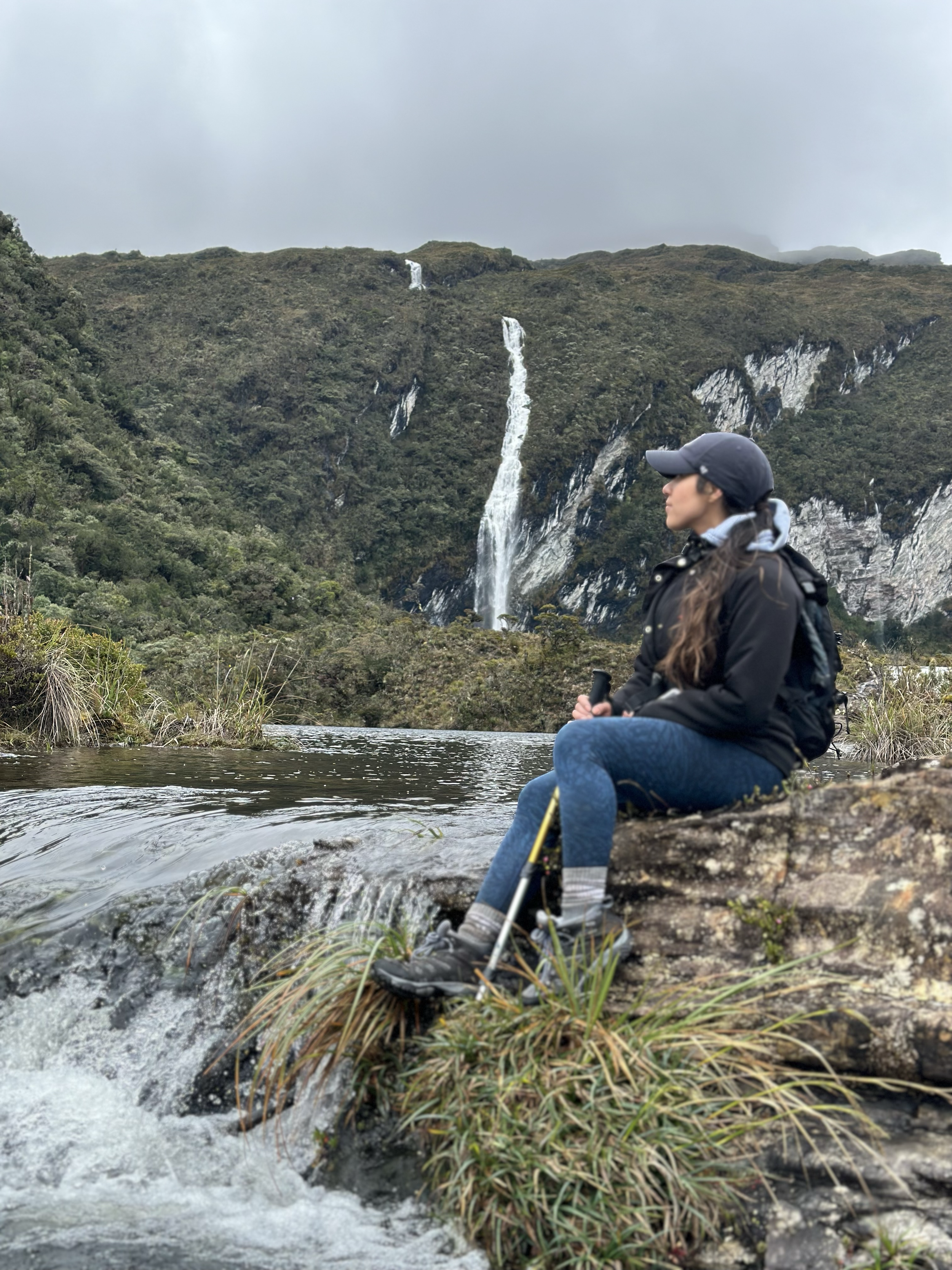 Mujer sentada en una roca junto a un río, con paisaje de bosque y cascadas de fondo.