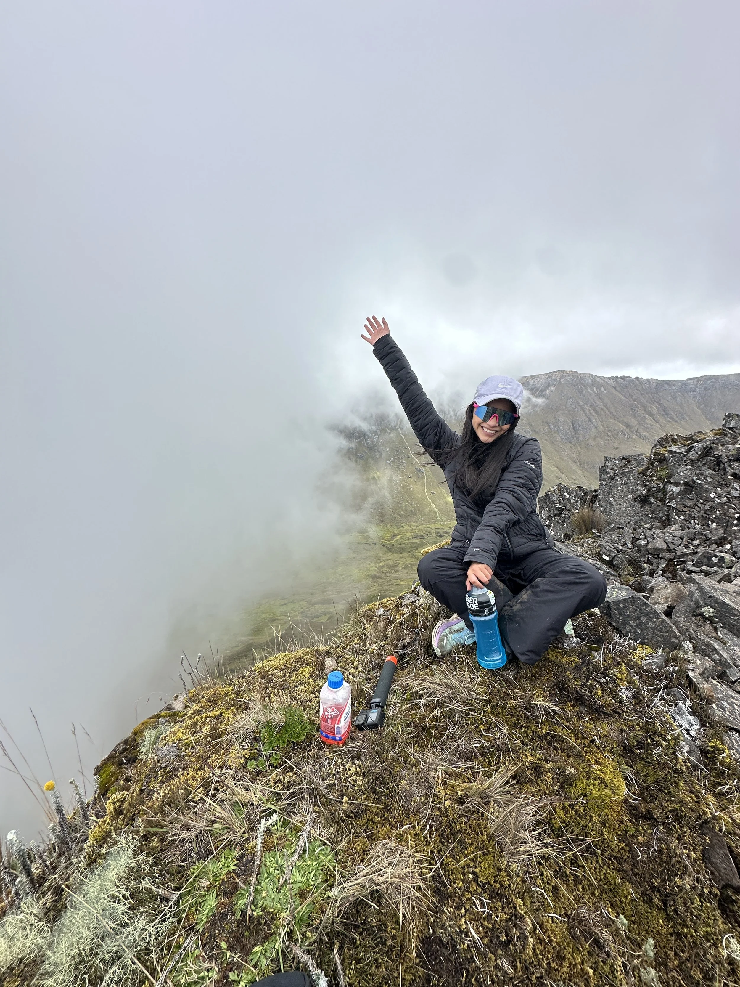 Mujer en ropa de abrigo en la montaña, rodeada de vegetación, con mochila y bebidas energéticas, sonriendo y levantando una mano, con nubes y niebla en el fondo.