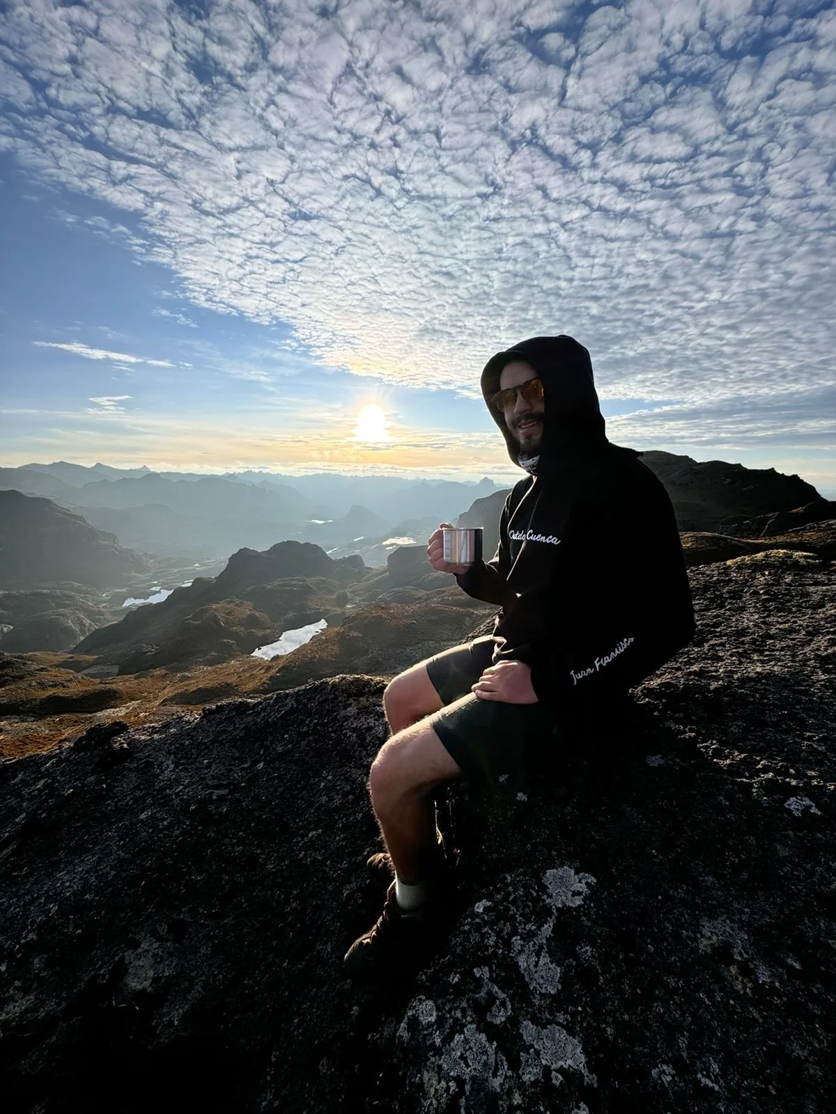 Hombre sentado en una roca en una montaña, sosteniendo una taza, con un paisaje de montañas y un cielo con nubes al amanecer.